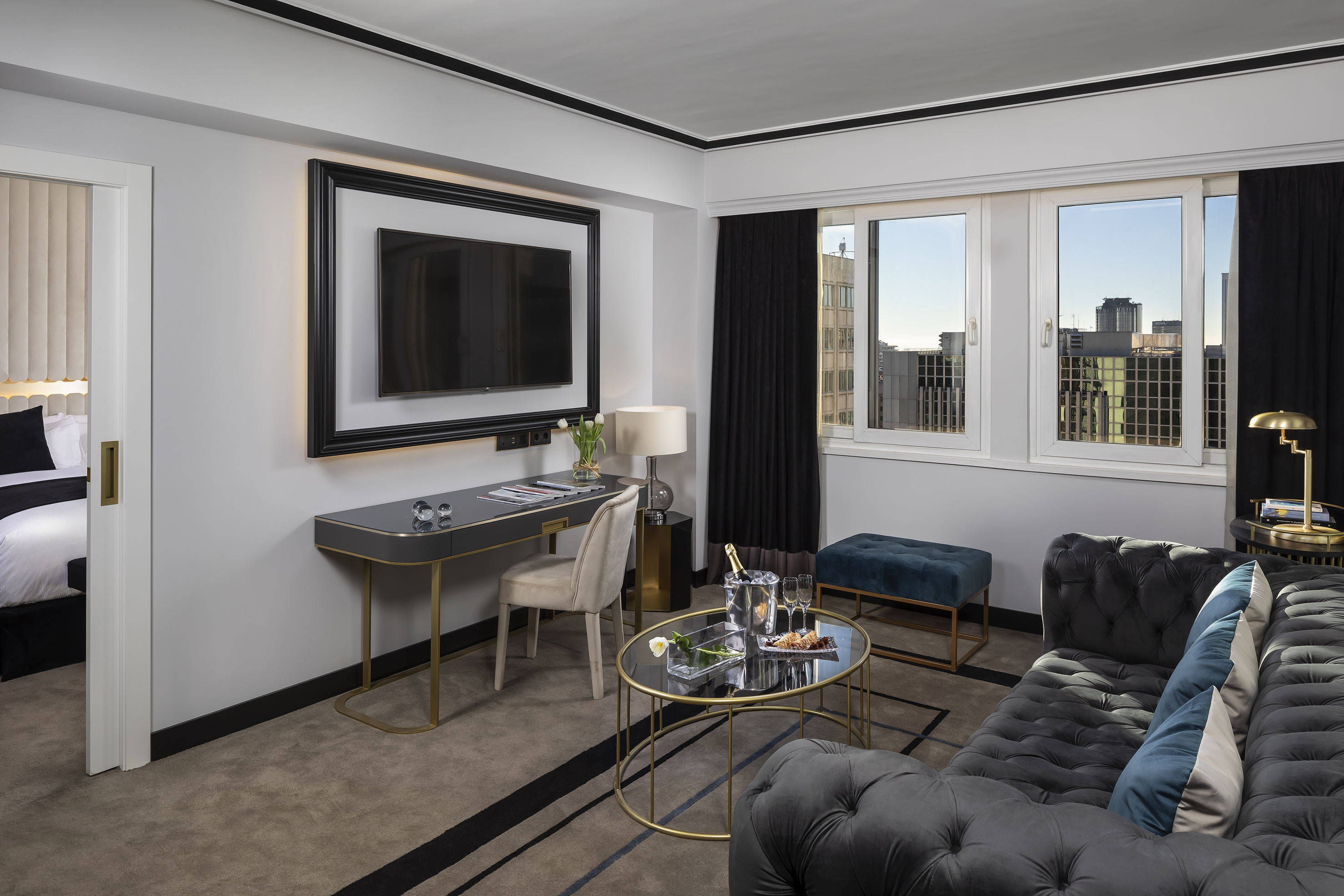 Luxury hotel suite living space with tufted sofa, desk, TV, city view.