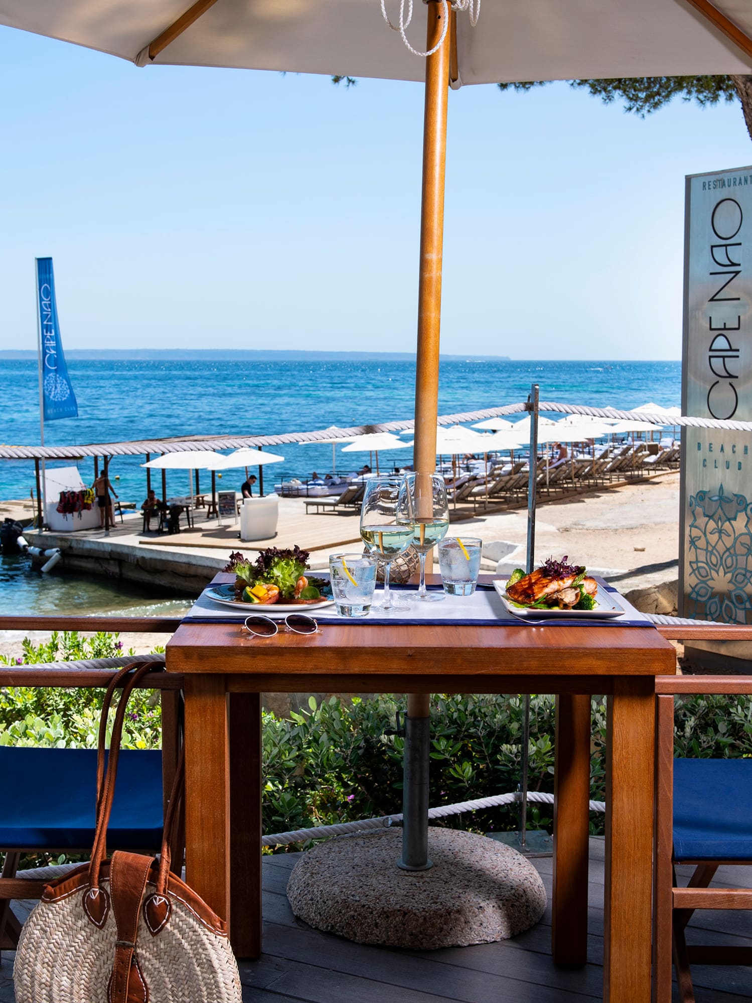 a table with food on it and chairs on a deck with a beach and water in the background