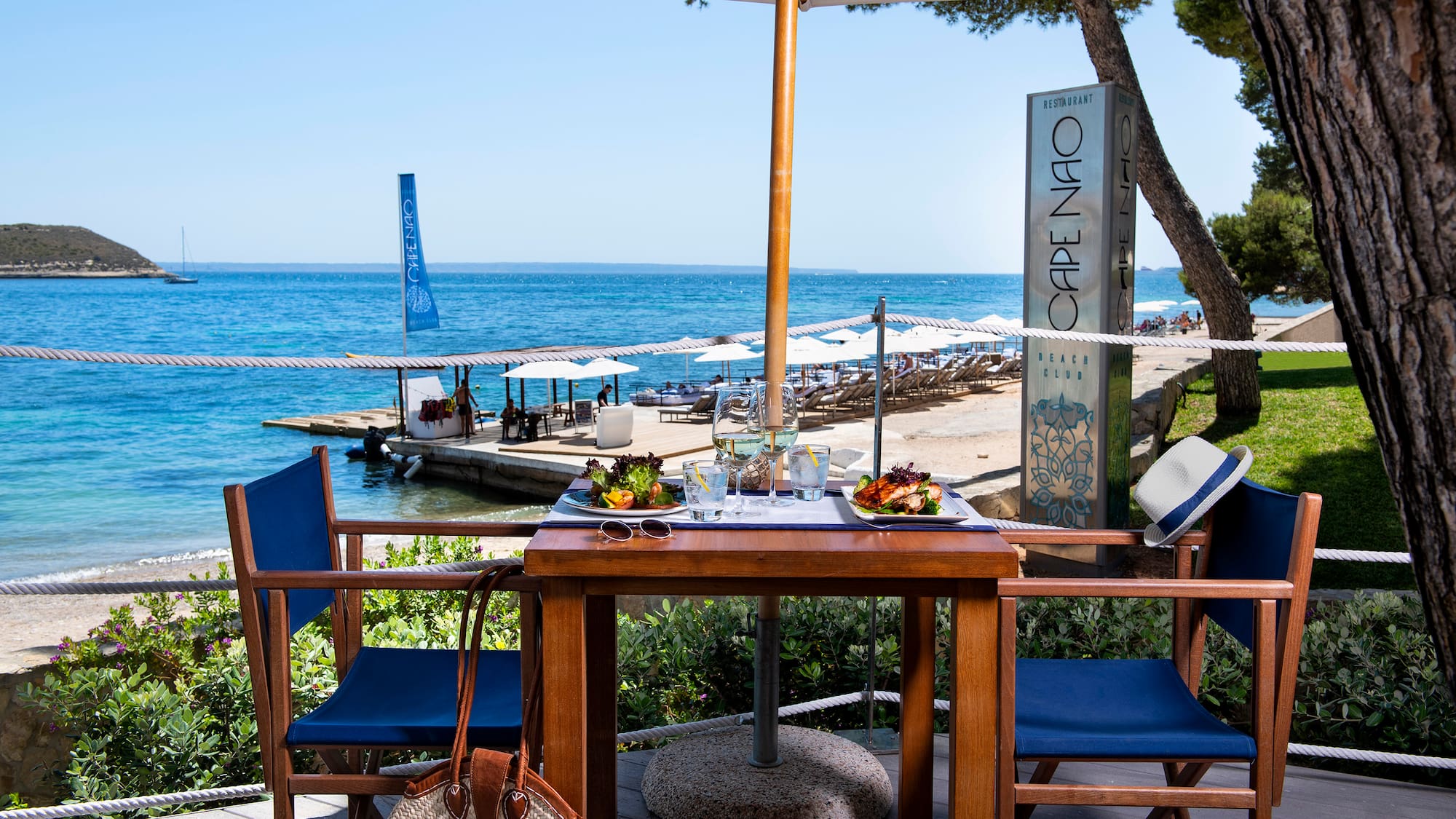 a table with food on it and chairs on a deck with a beach and water in the background