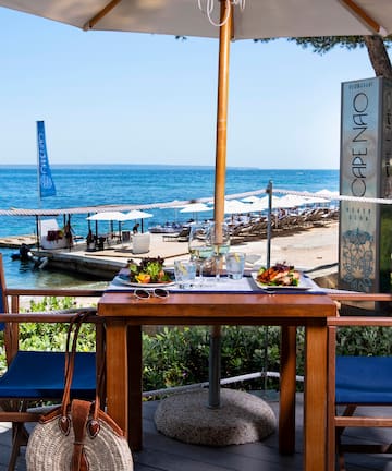 a table with food on it and chairs on a deck with a beach and water in the background