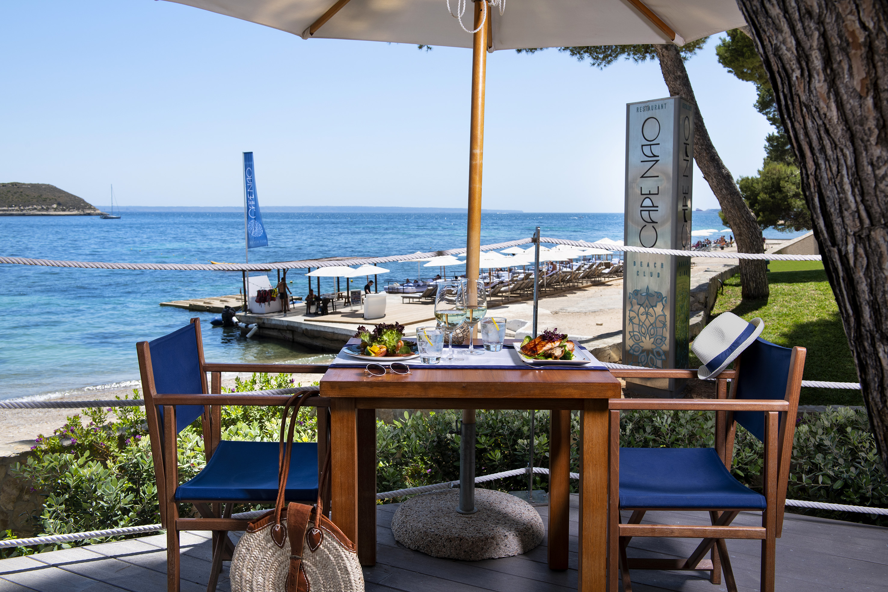 a table with food on it and chairs on a deck with a beach and water in the background