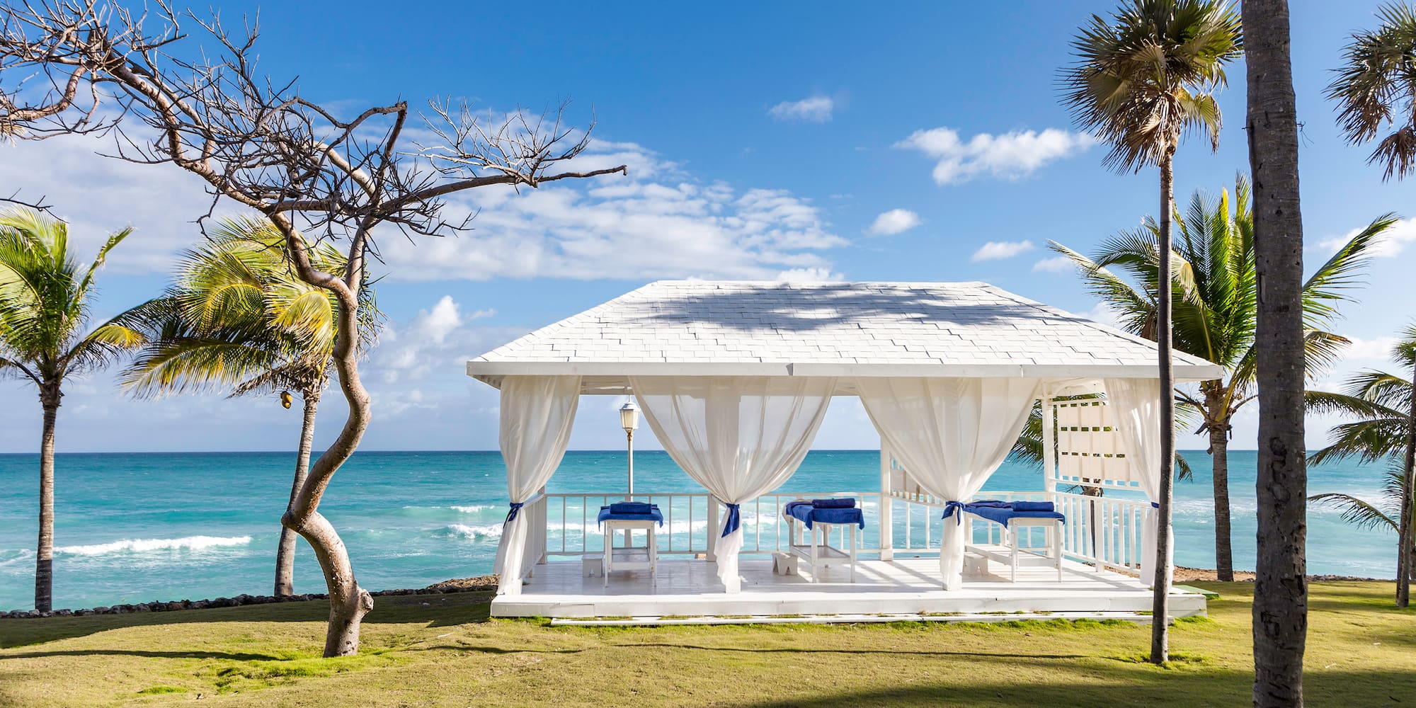 a white gazebo with blue and white curtains and a beach in the background