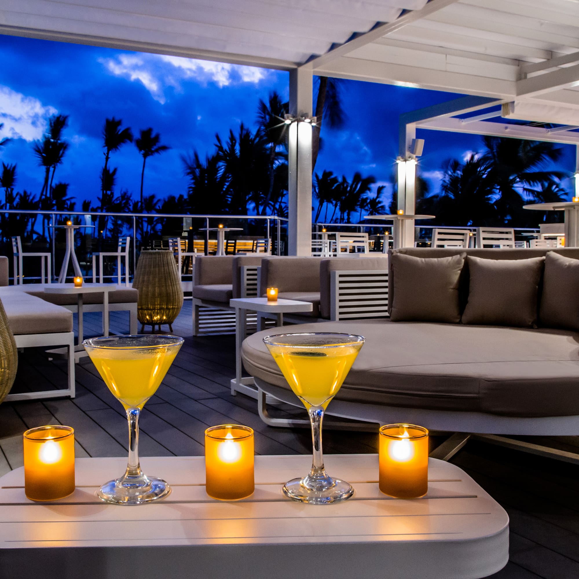 Tropical outdoor lounge with cocktails, glowing candles, and palm trees at dusk.