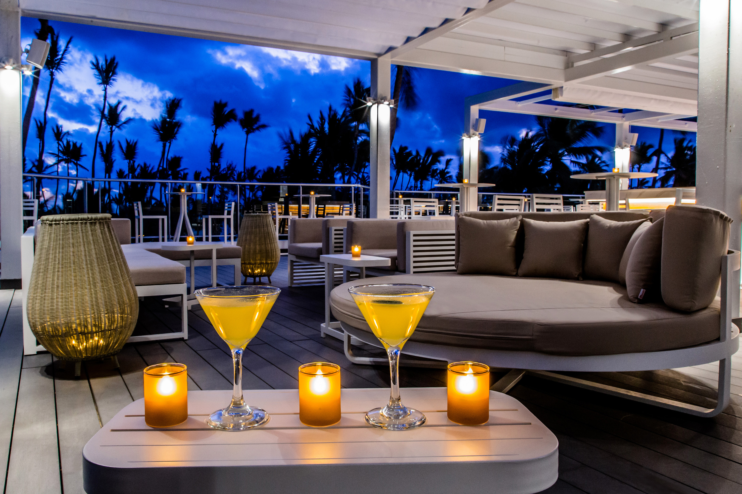 Tropical outdoor lounge with cocktails, glowing candles, and palm trees at dusk.