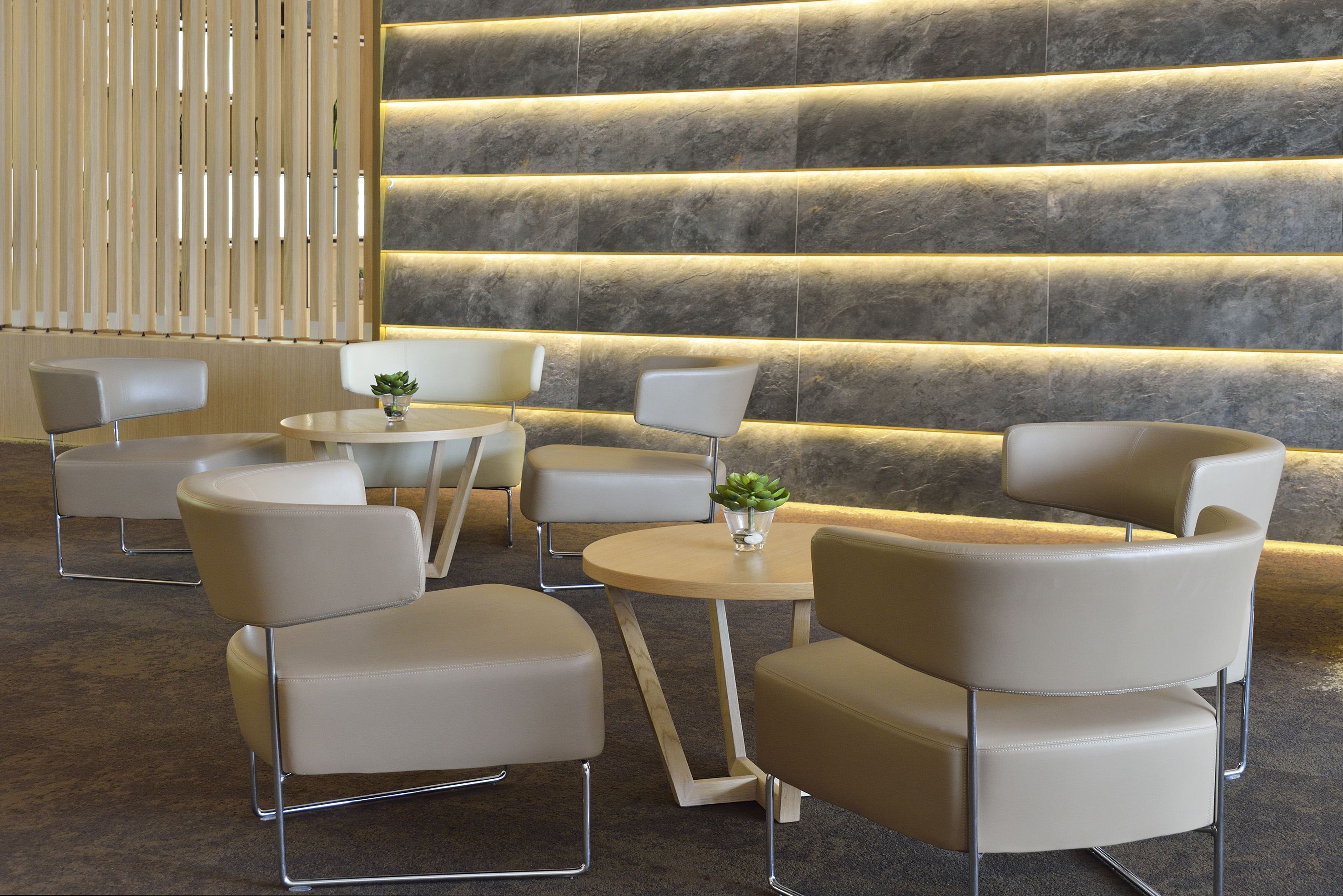 Contemporary lounge with beige armchairs, wood tables, and backlit stone wall.
