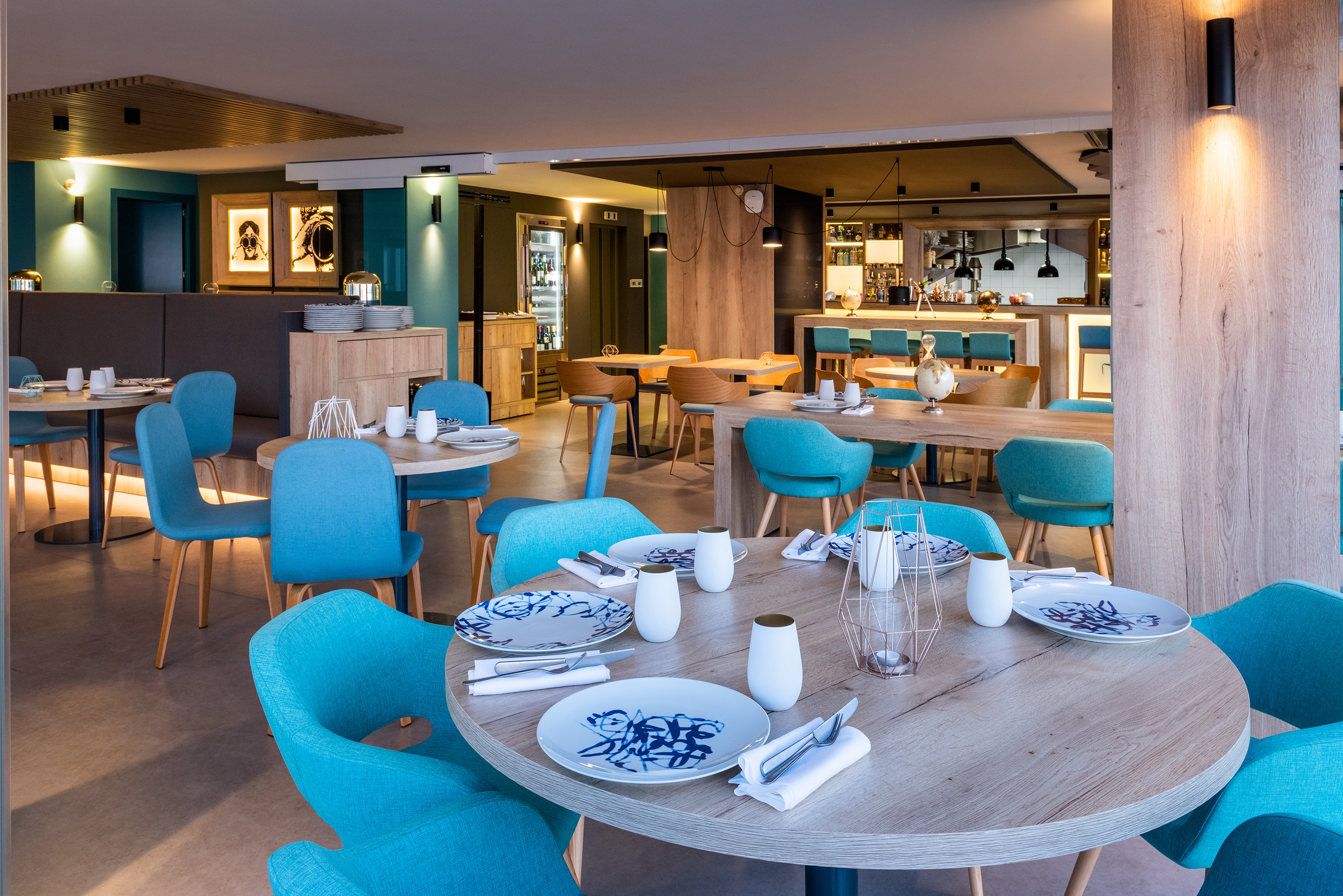 Modern restaurant interior with teal dining chairs, wooden tables, and bar area.