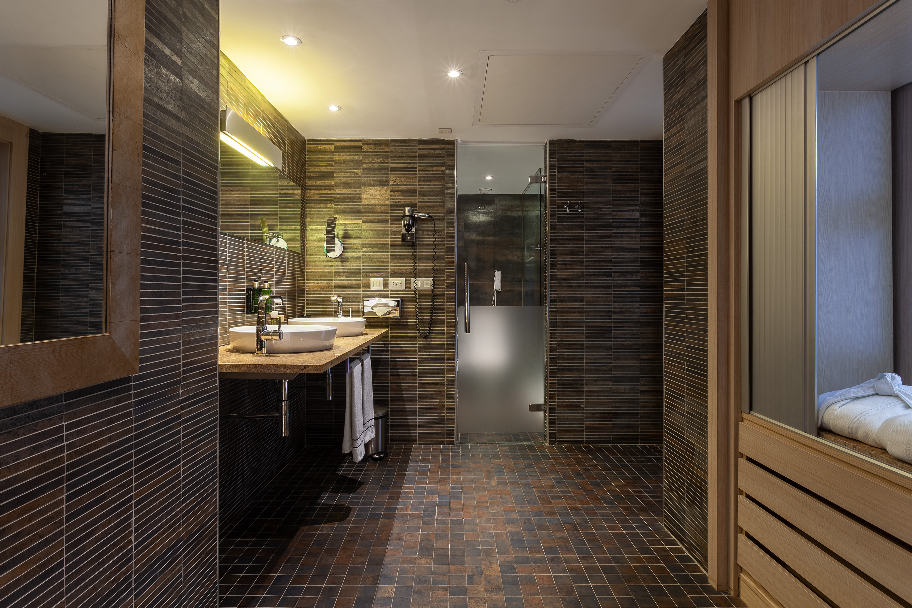 Modern hotel bathroom with dark tiles, double vanity, glass shower.