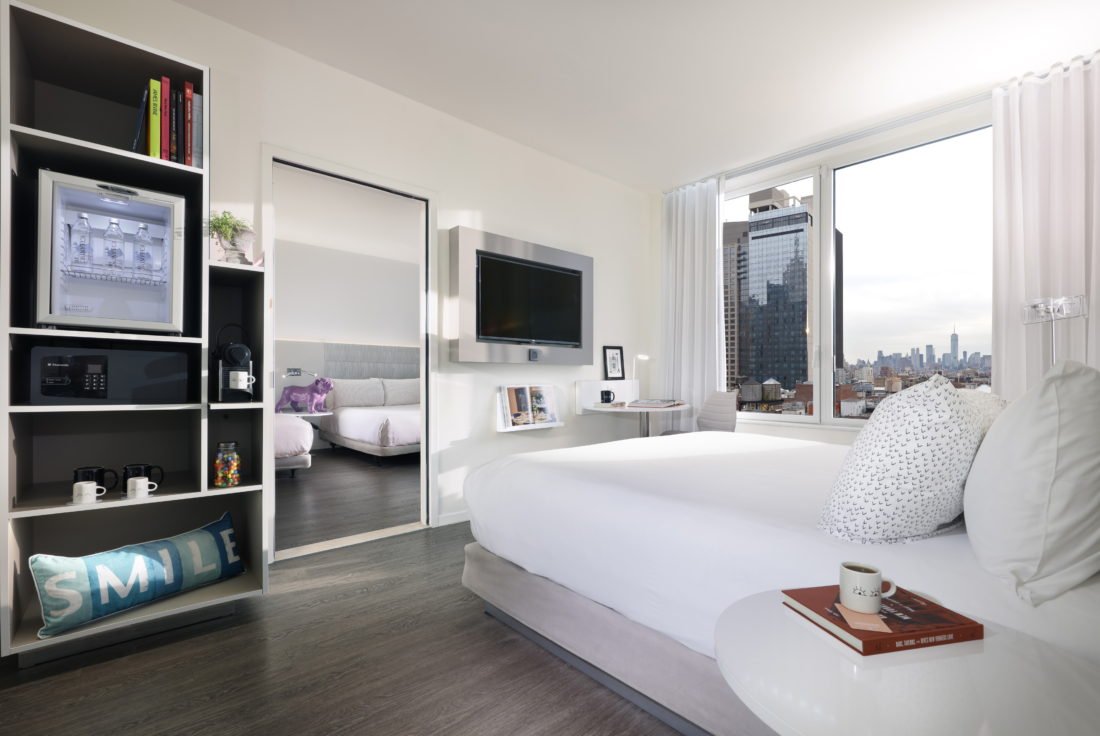 Modern hotel room with NYC skyline view, large bed, flat-screen TV, and mini-fridge.