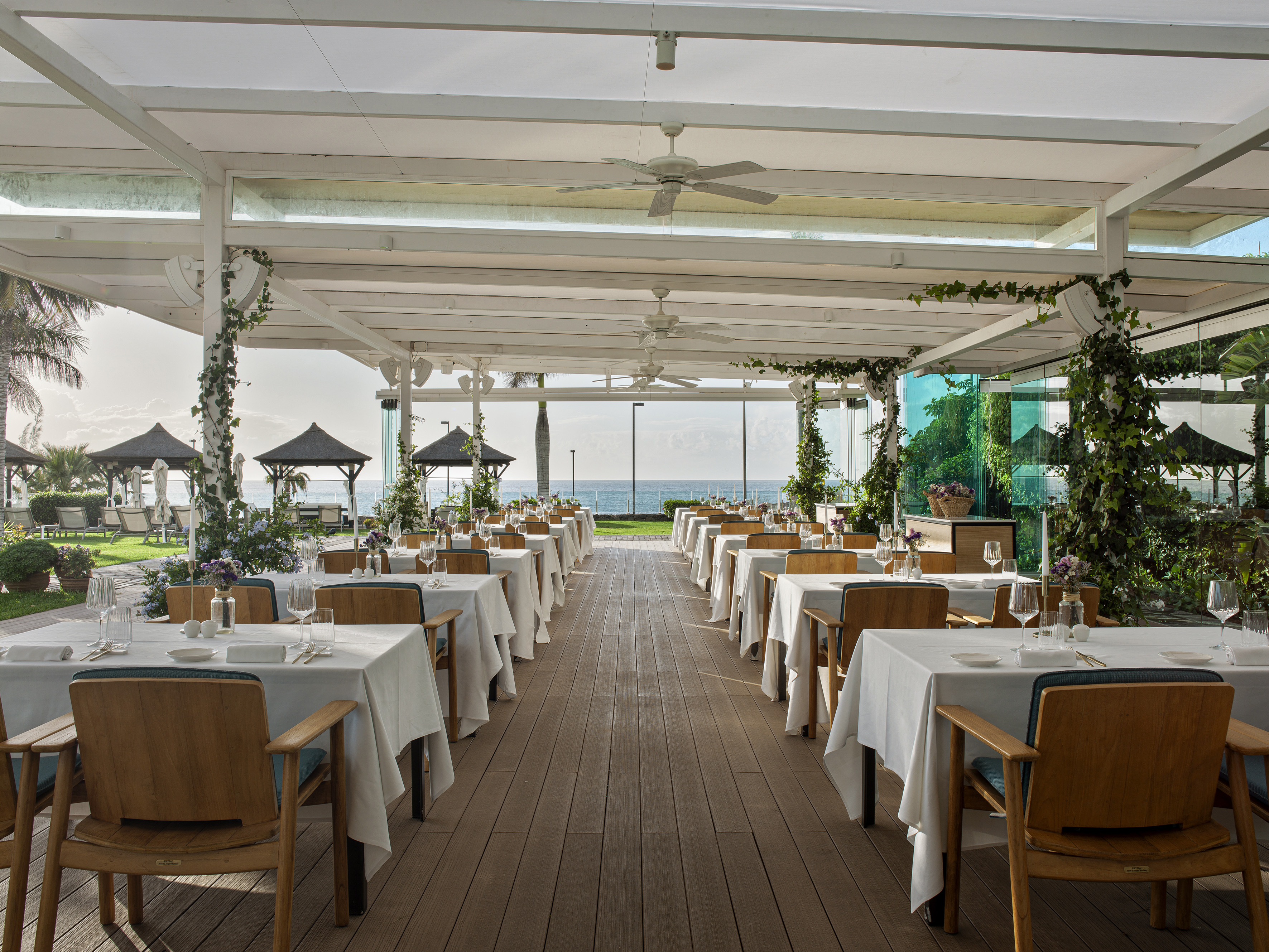 Outdoor restaurant patio with sea view, set tables, and wooden dining chairs.