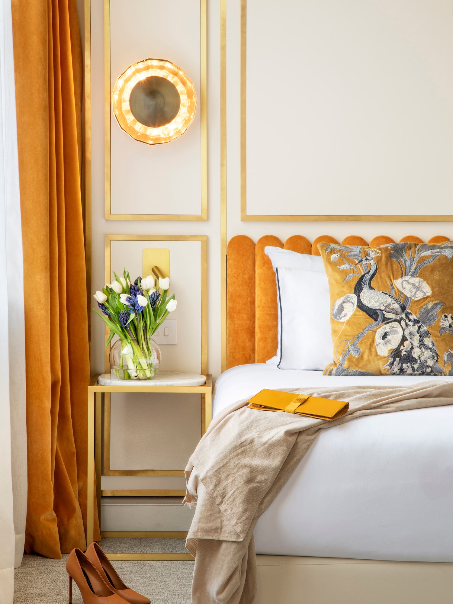 Luxe hotel room: mustard velvet headboard, peacock pillow, gold accents, flowers.