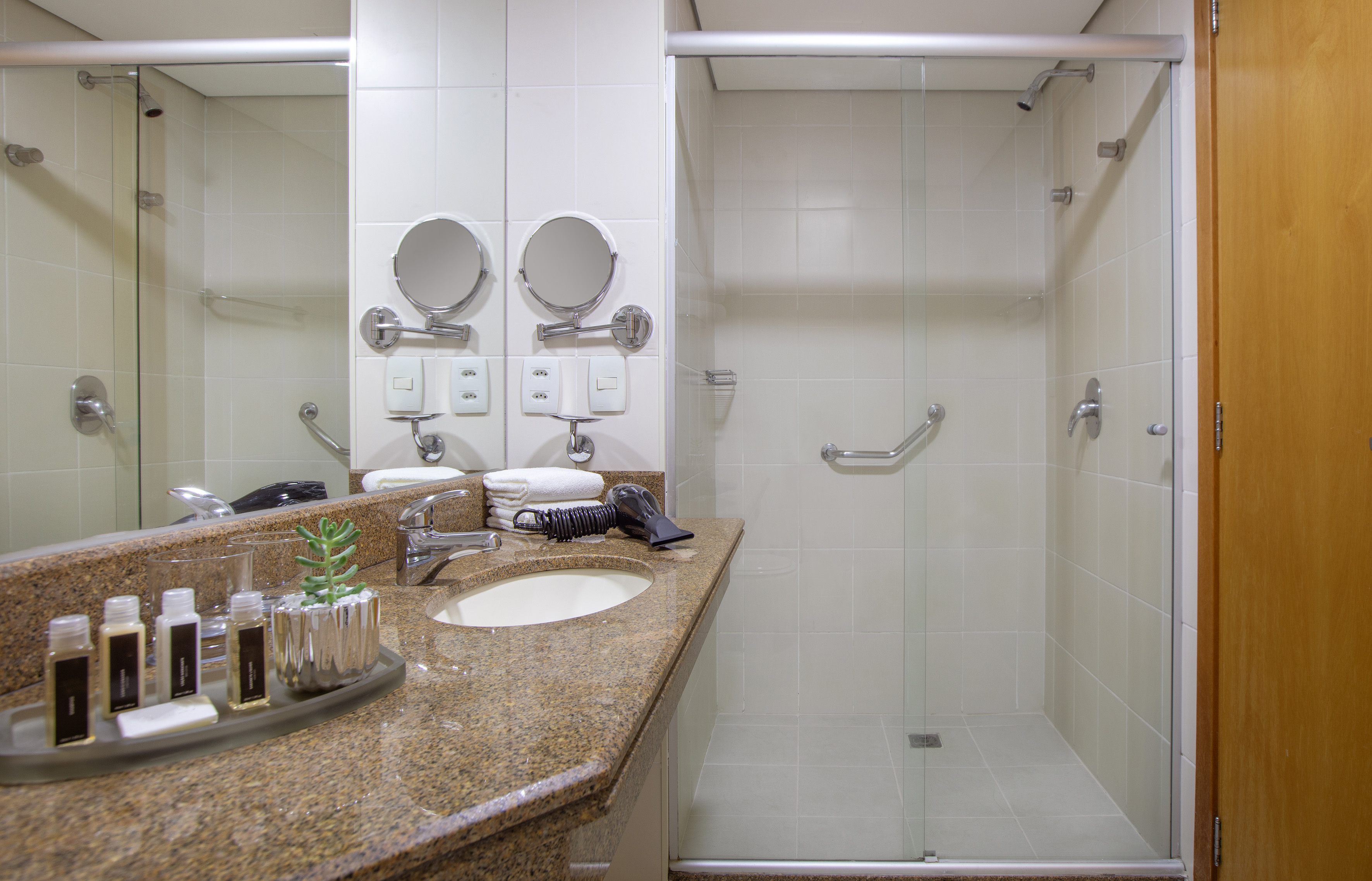 Hotel bathroom with granite vanity, sink, magnifying mirrors, and walk-in shower.