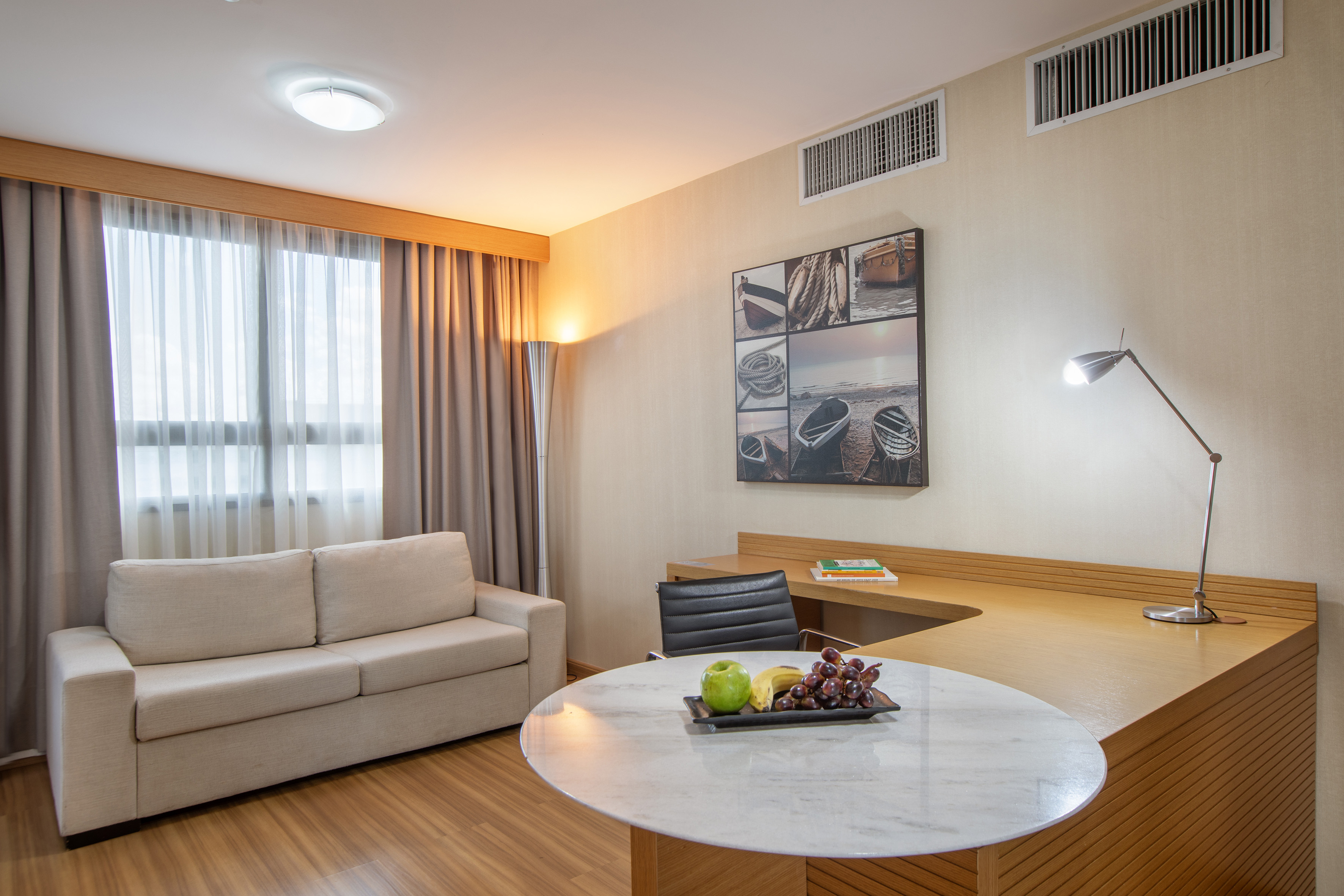 Modern hotel suite living area with sofa, desk, fruit table, and nautical art.