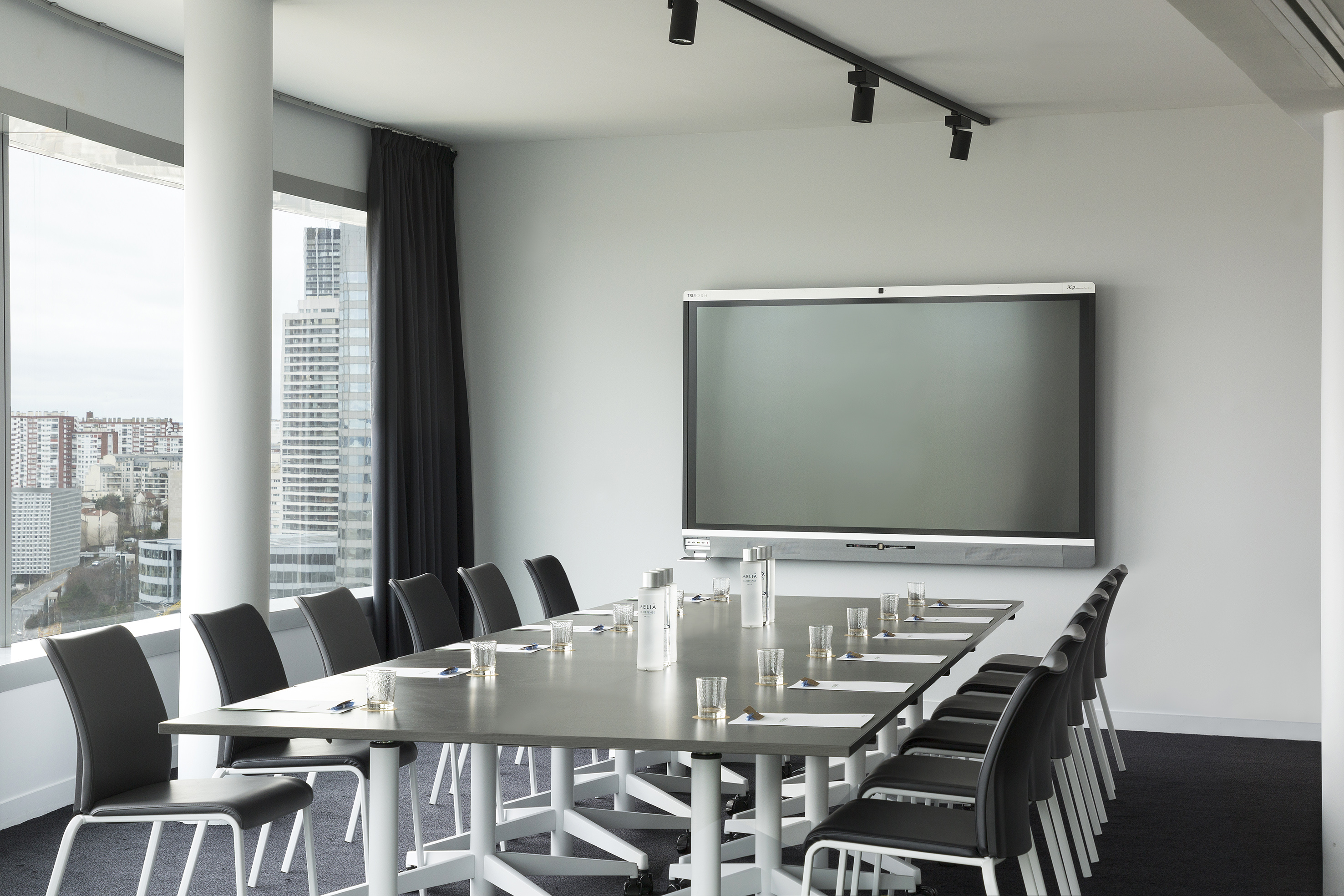 Sleek corporate meeting room with large display, long table, chairs, and city view.