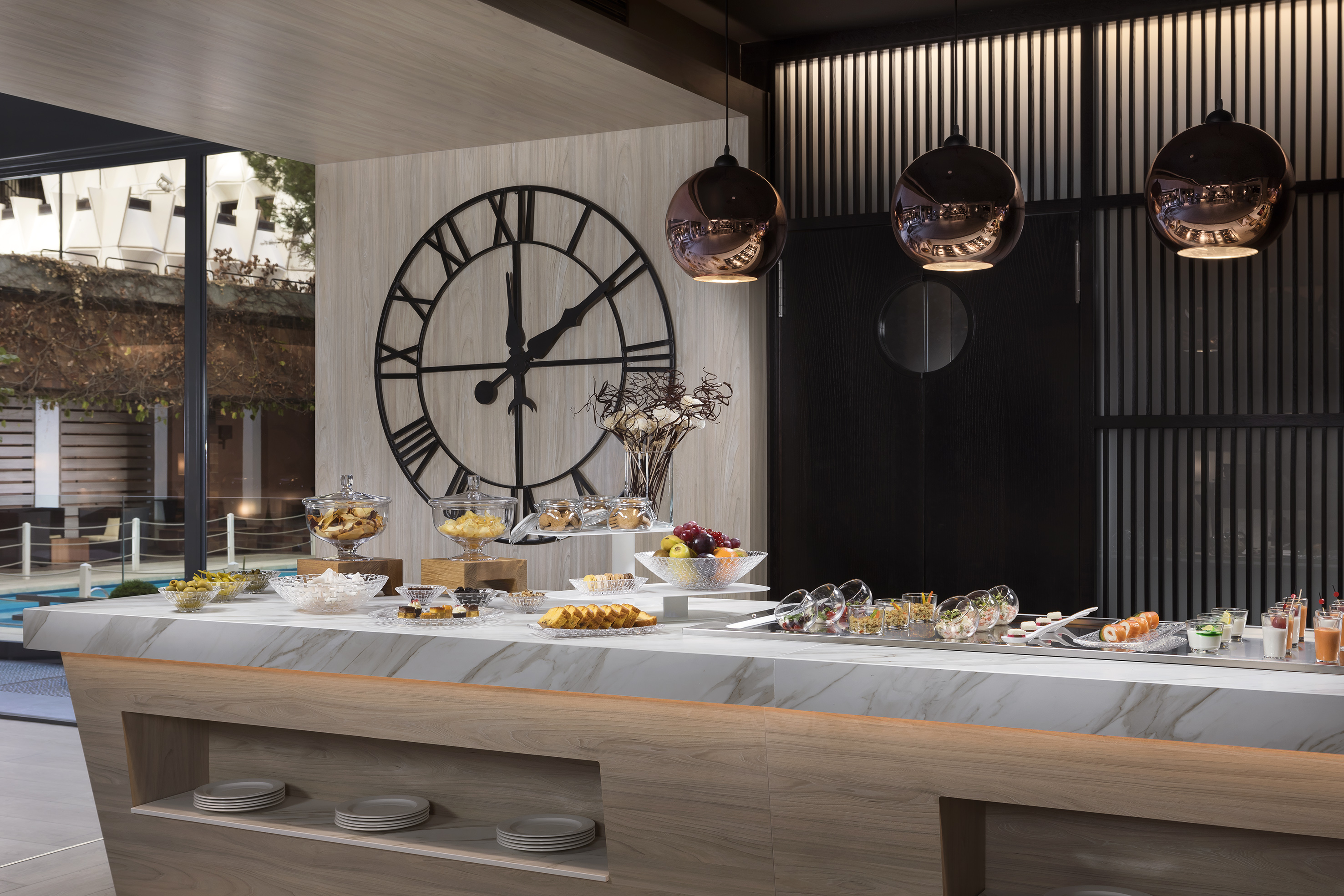 Modern hotel buffet with diverse breakfast spread, snacks, fruits, and large clock.