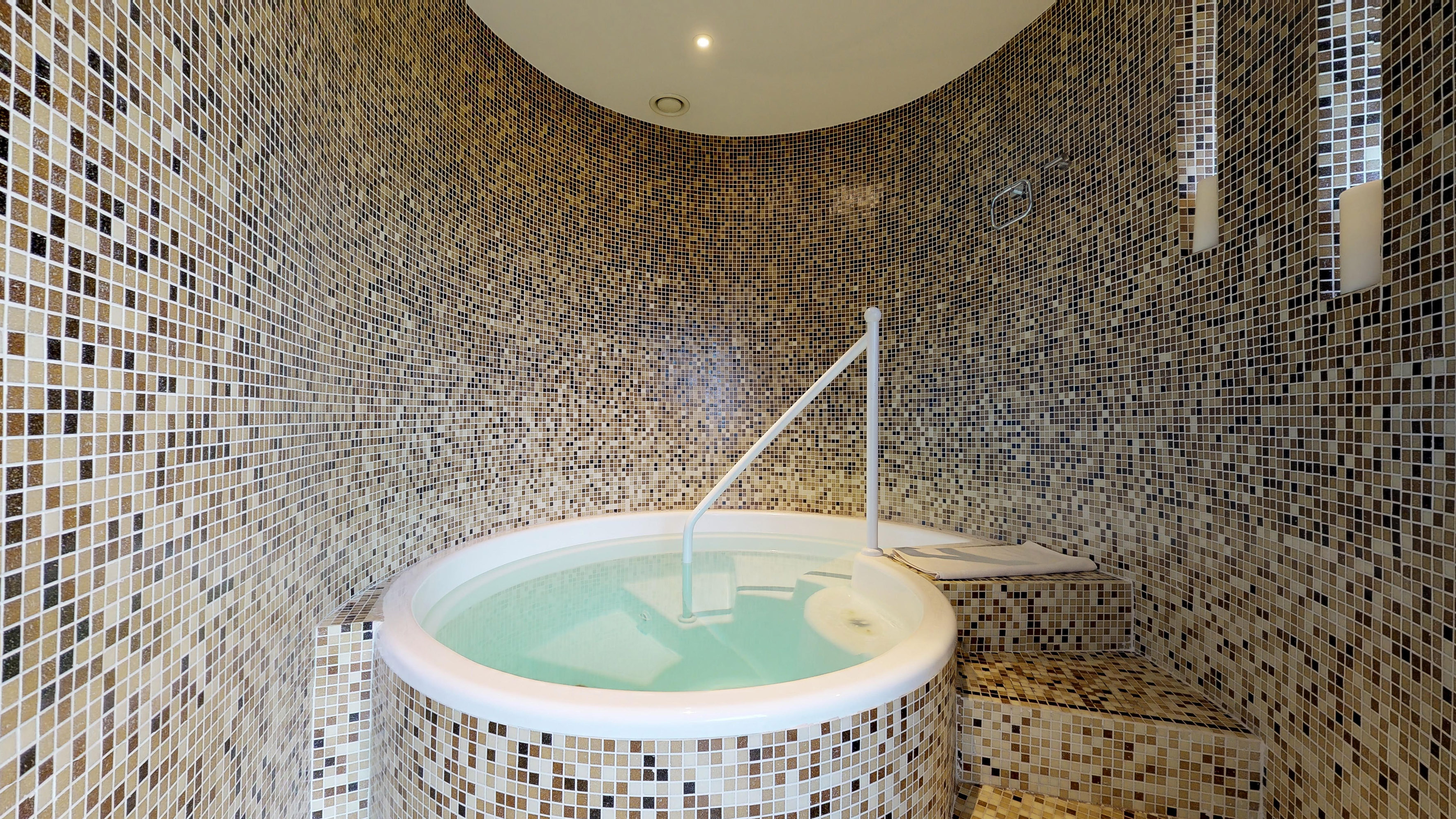 Relaxing mosaic tiled hot tub with light blue water and handrail.