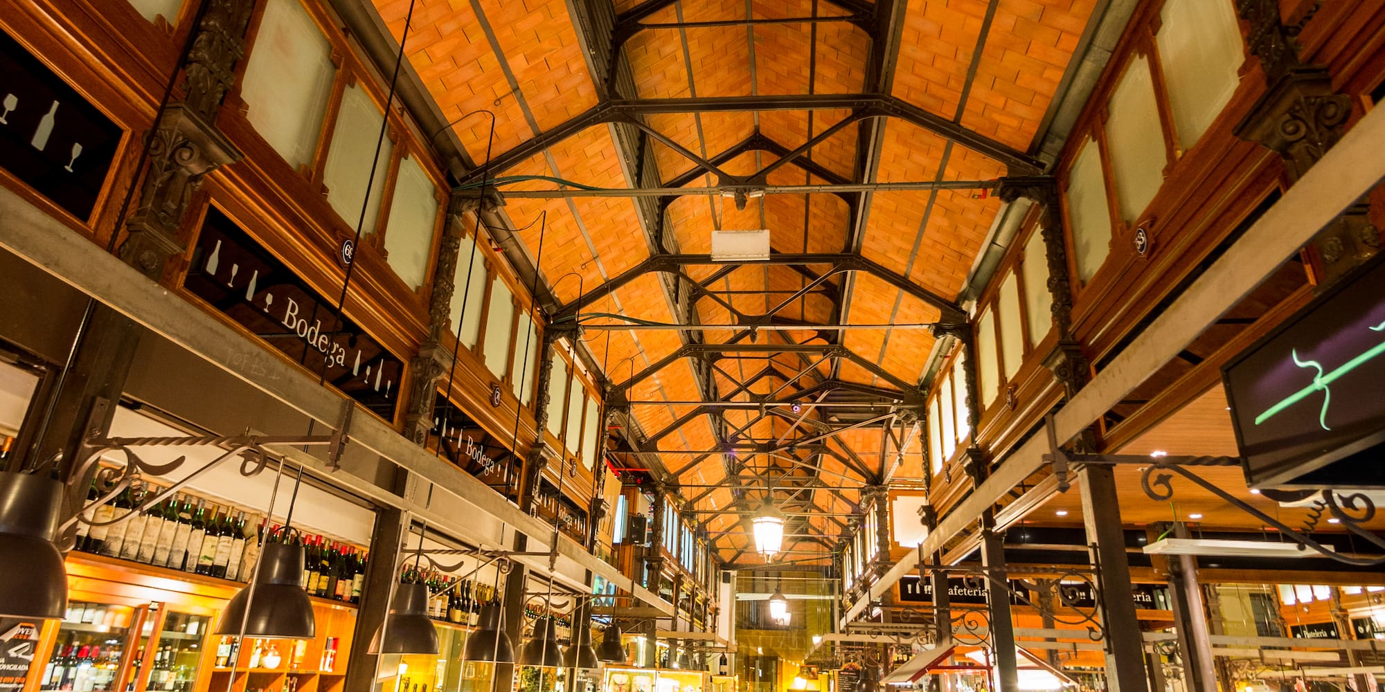 Mercado español animado, gente en puestos de tapas y bebidas bajo techos de teja y metal.