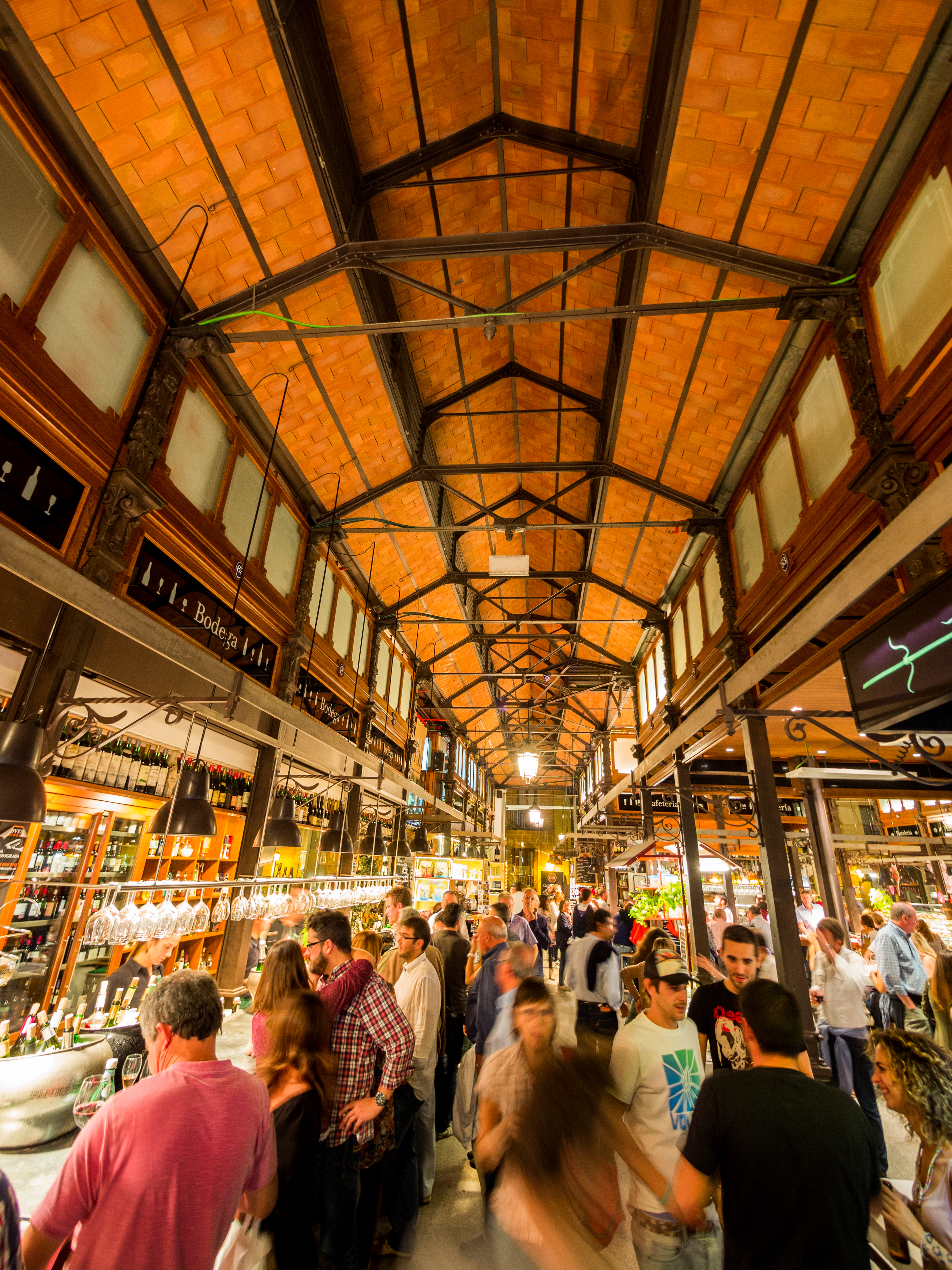 Mercado español animado, gente en puestos de tapas y bebidas bajo techos de teja y metal.