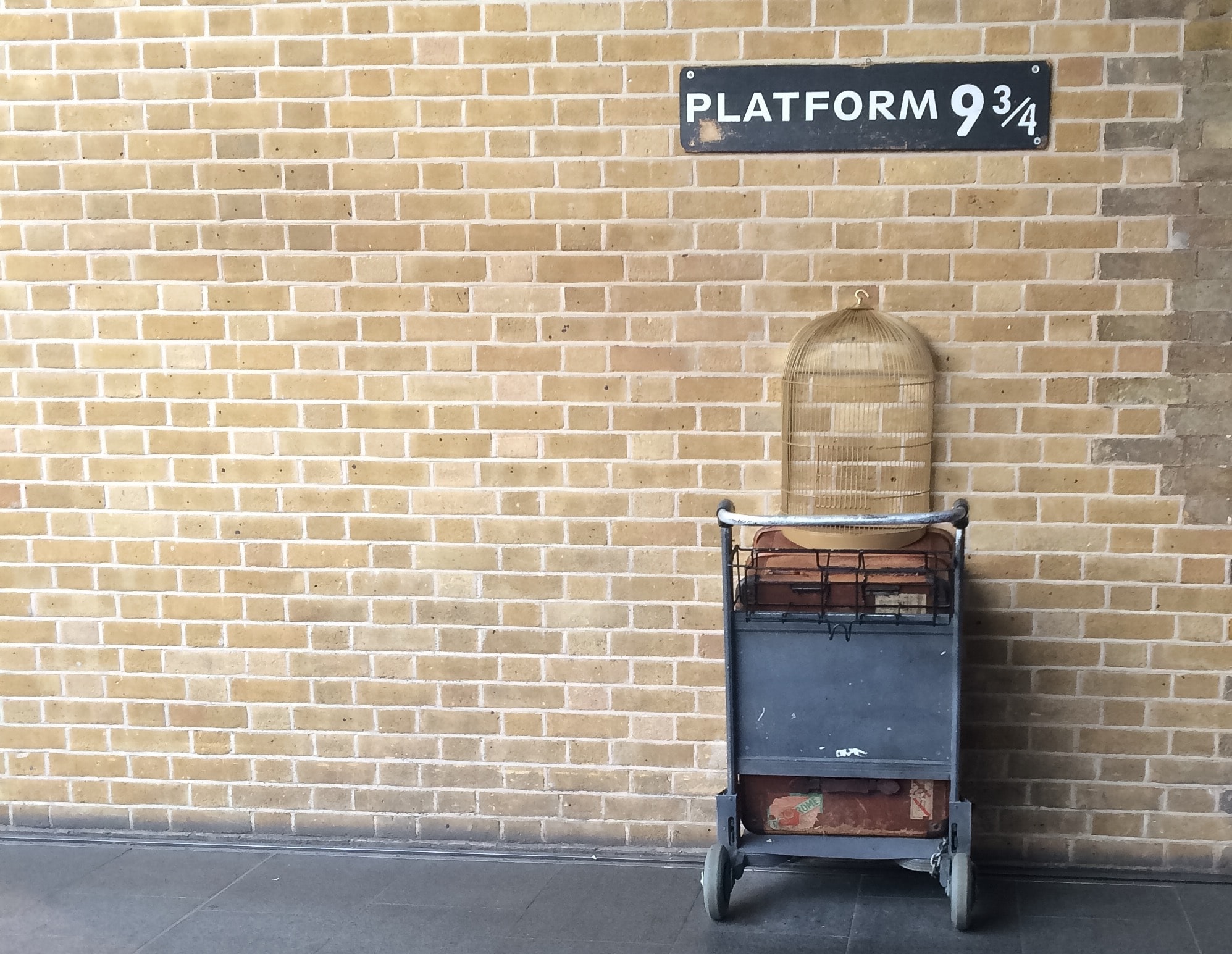 Platform 9 3/4 sign and luggage trolley with birdcage on brick wall.