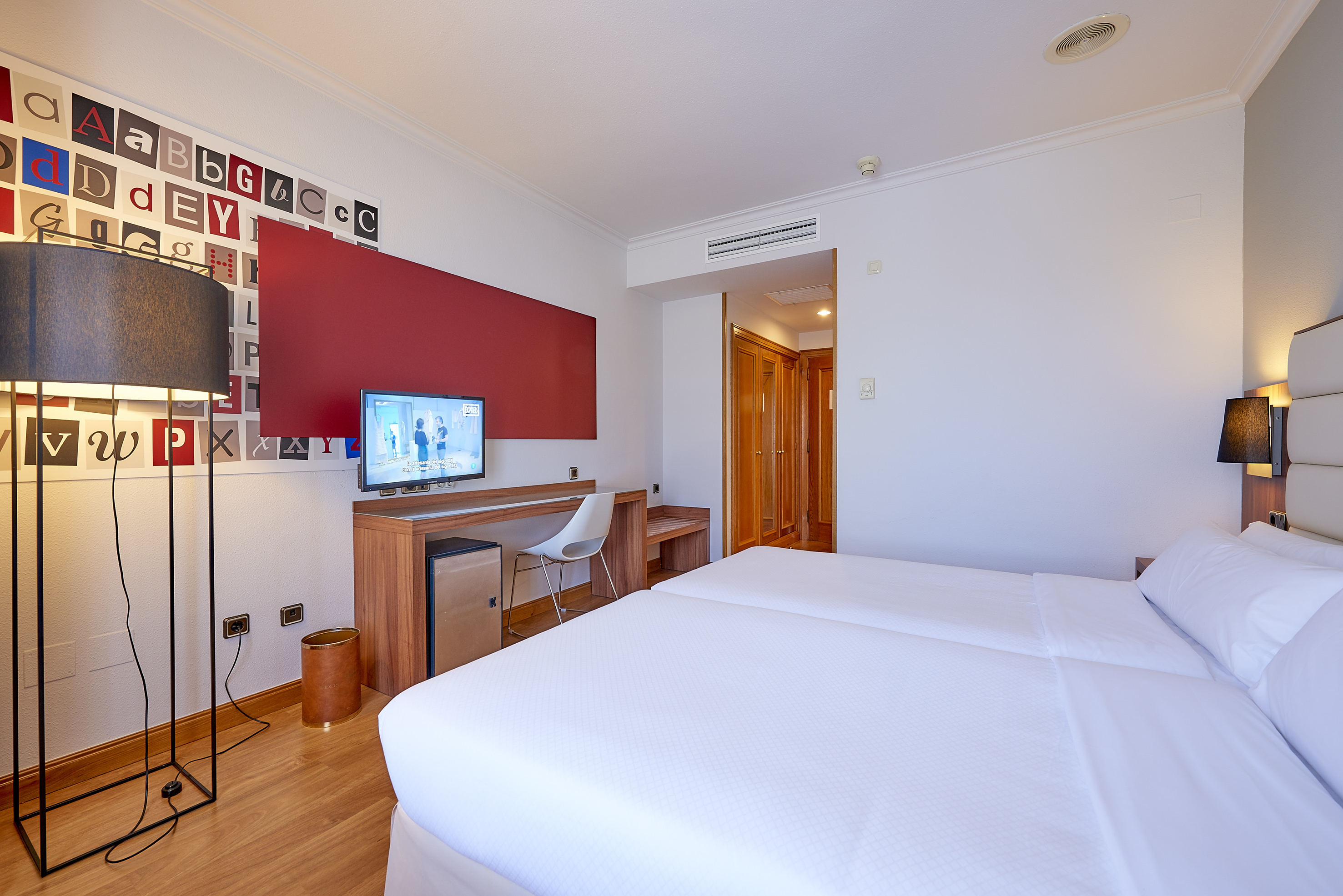 Modern hotel room with twin beds, desk, TV, and alphabet wall decor.