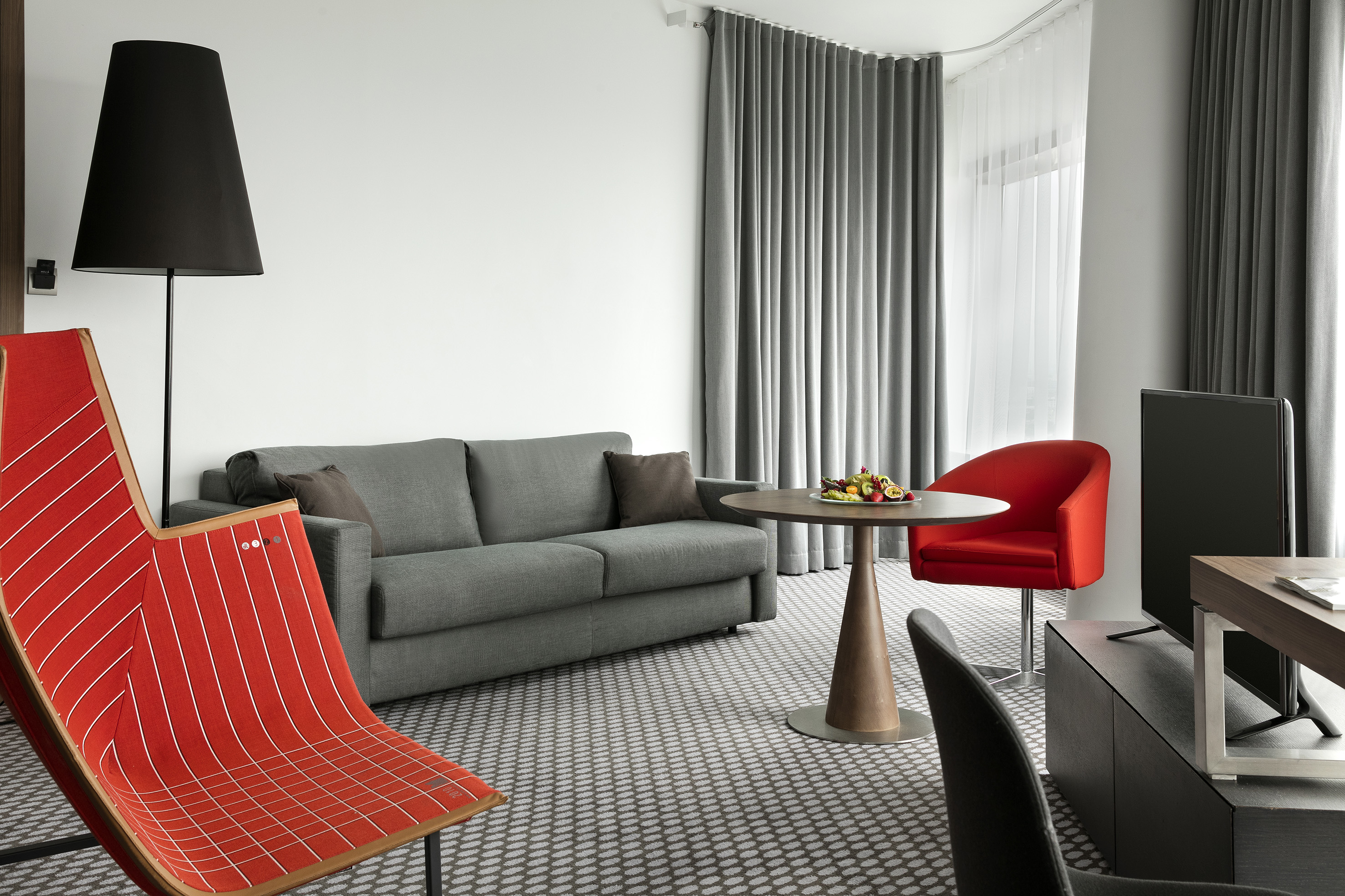 Modern hotel living room with grey sofa, red chairs, table, and TV.