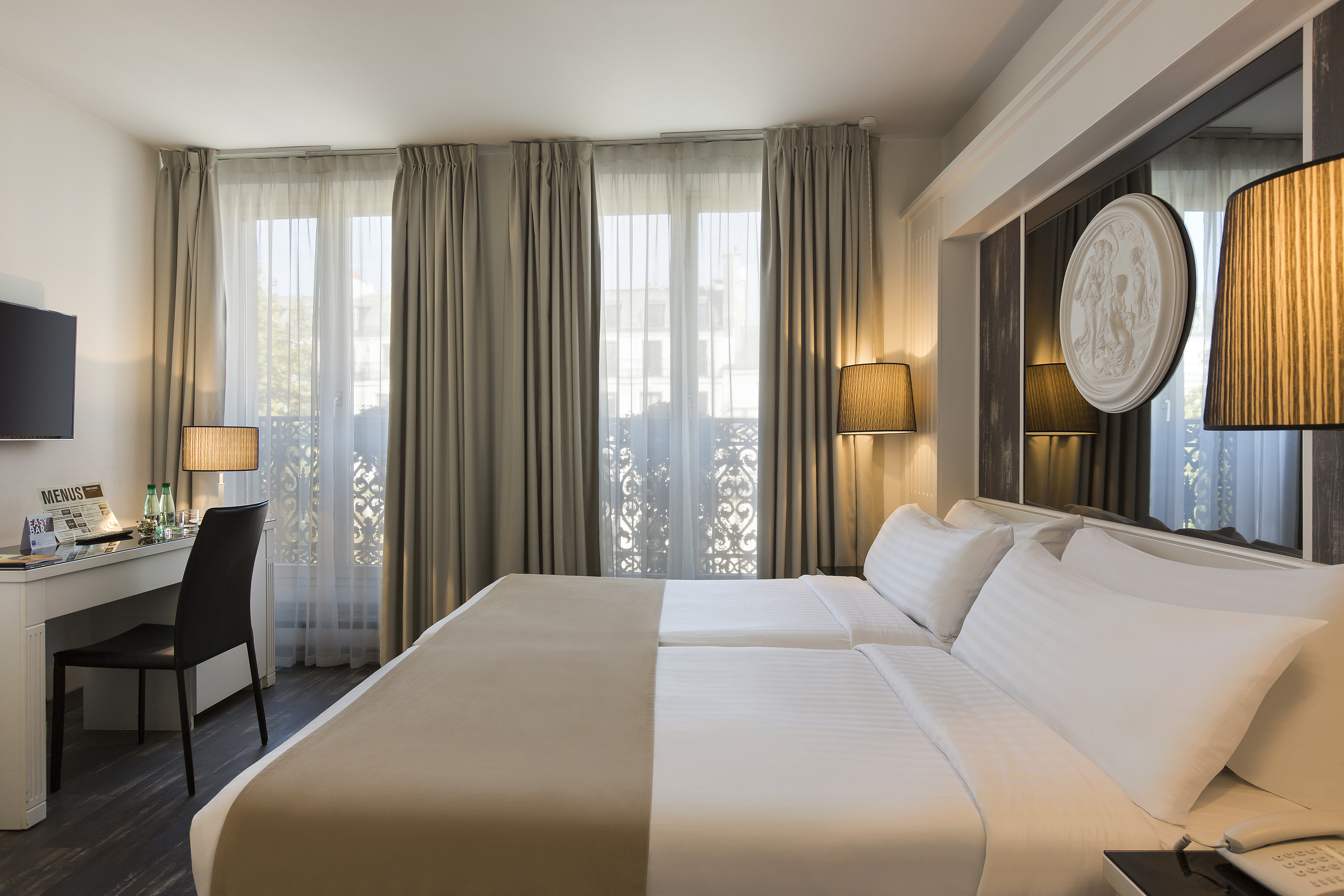 Elegant hotel room with twin beds, modern desk, and large window with ornate balcony.