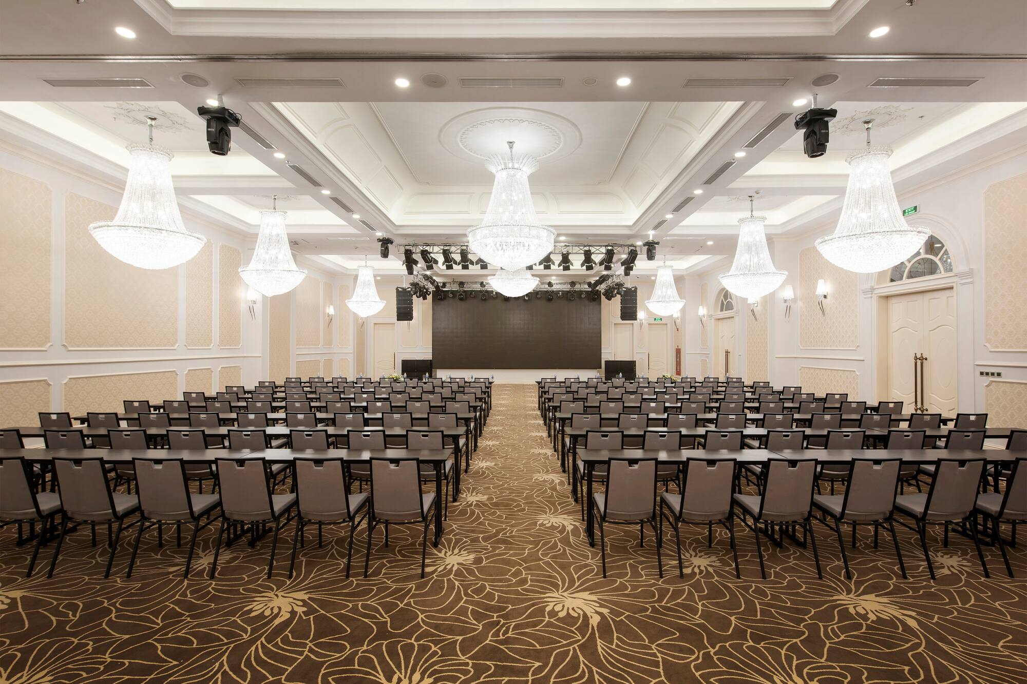 Spacious, elegant conference room with tables, chairs, stage, and chandeliers.