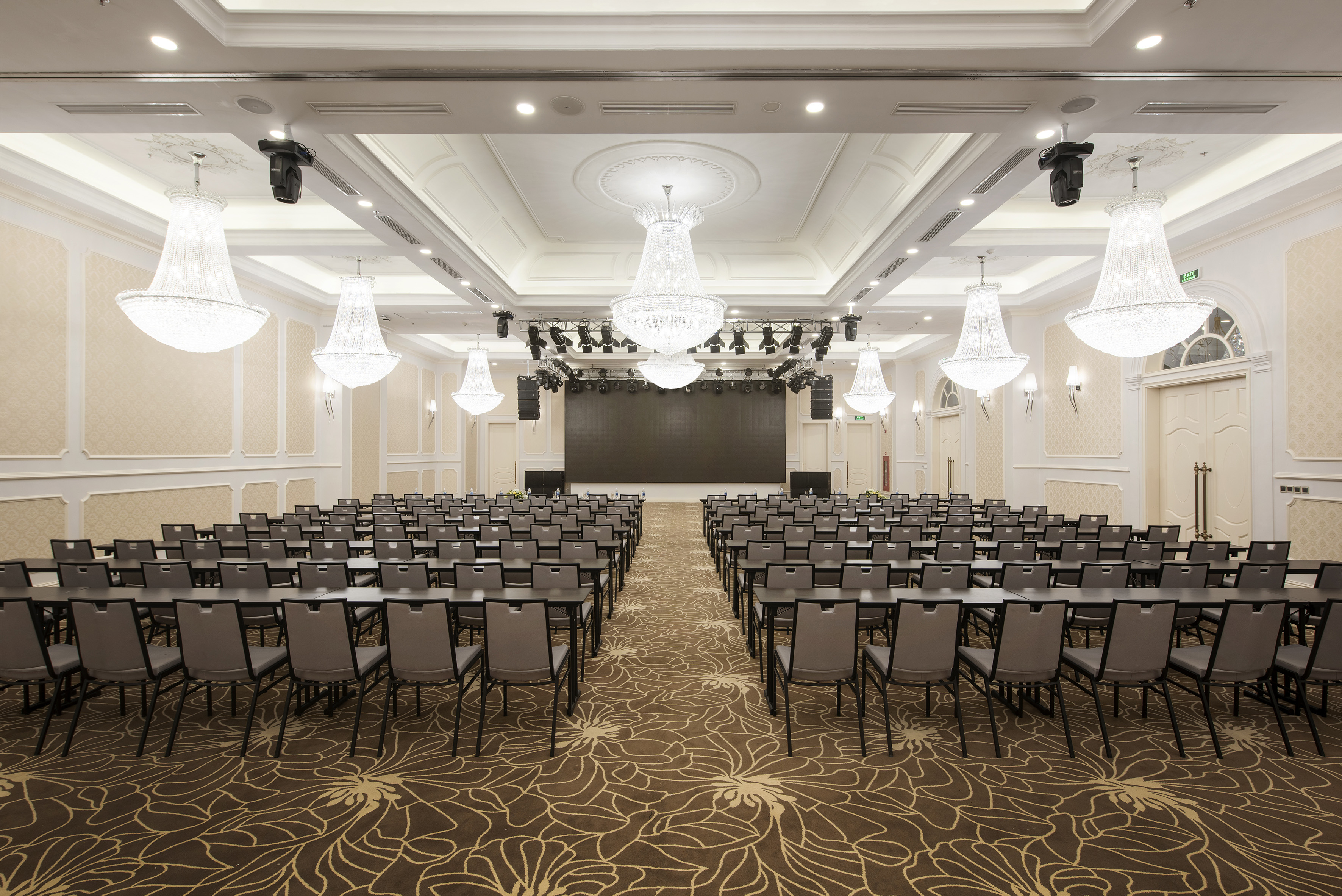 Spacious, elegant conference room with tables, chairs, stage, and chandeliers.