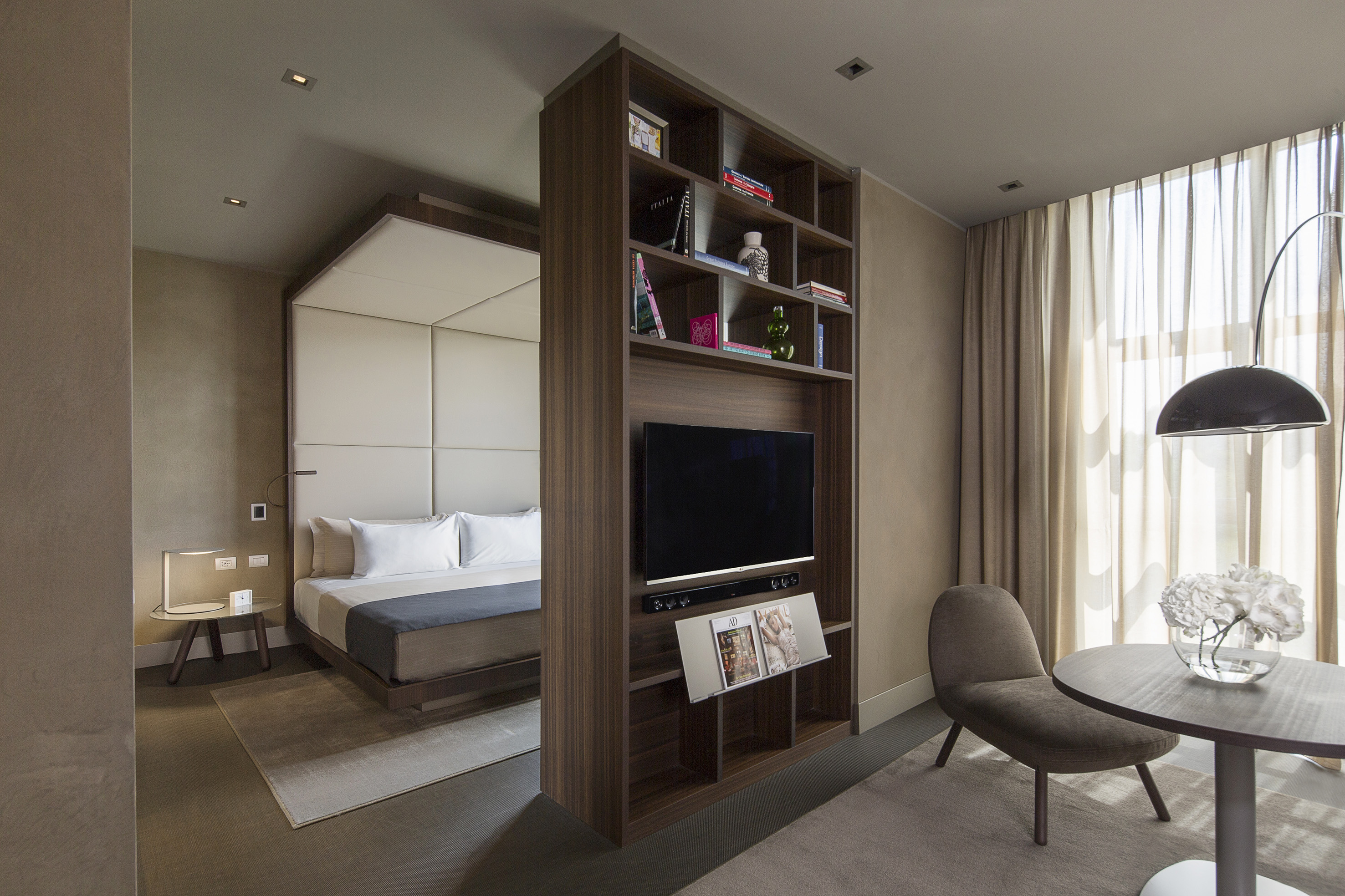 Modern hotel suite featuring bed, TV bookshelf divider, and neutral sitting area.
