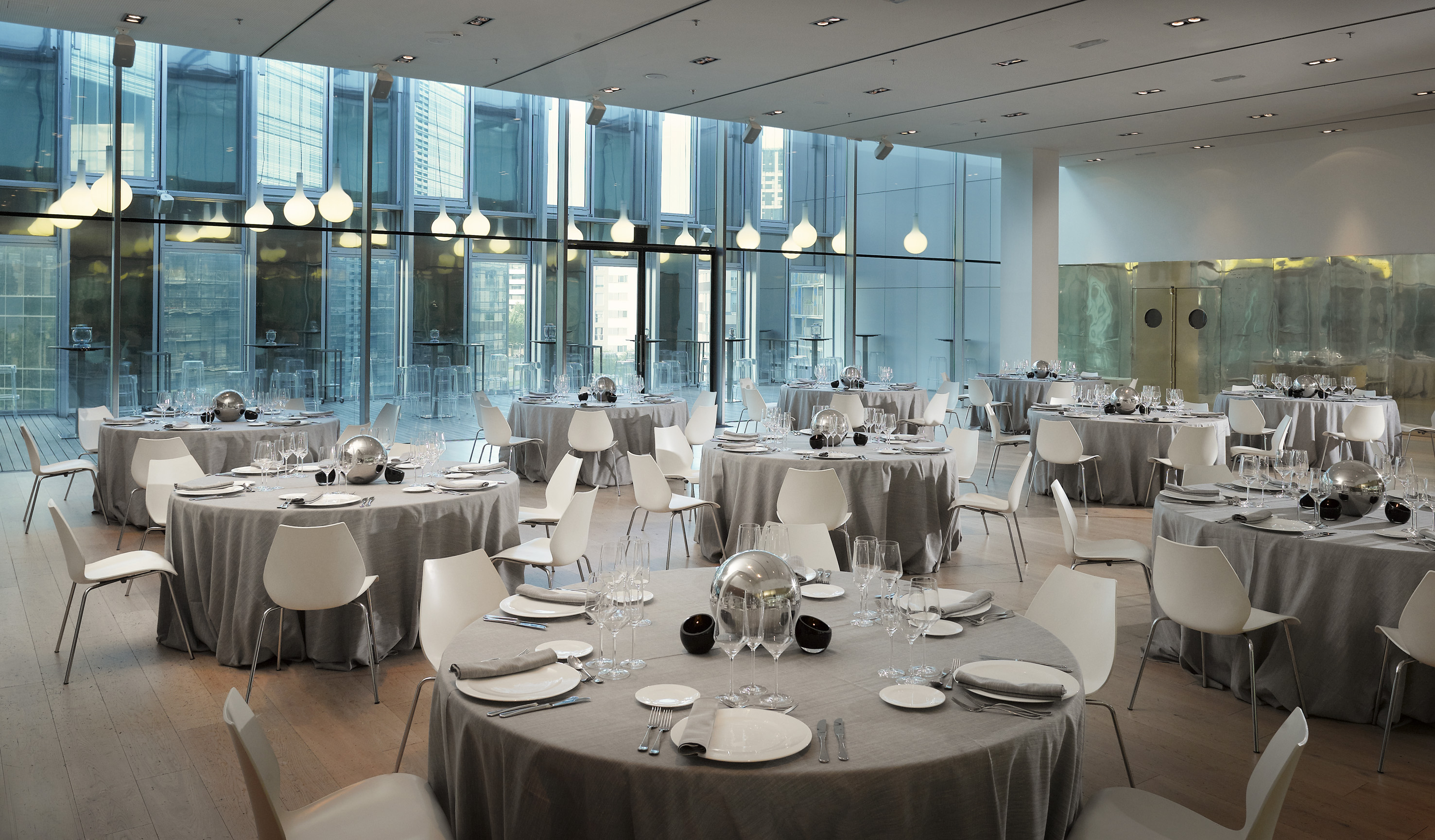 Modern banquet hall, round tables, grey linens, white chairs, large glass windows.
