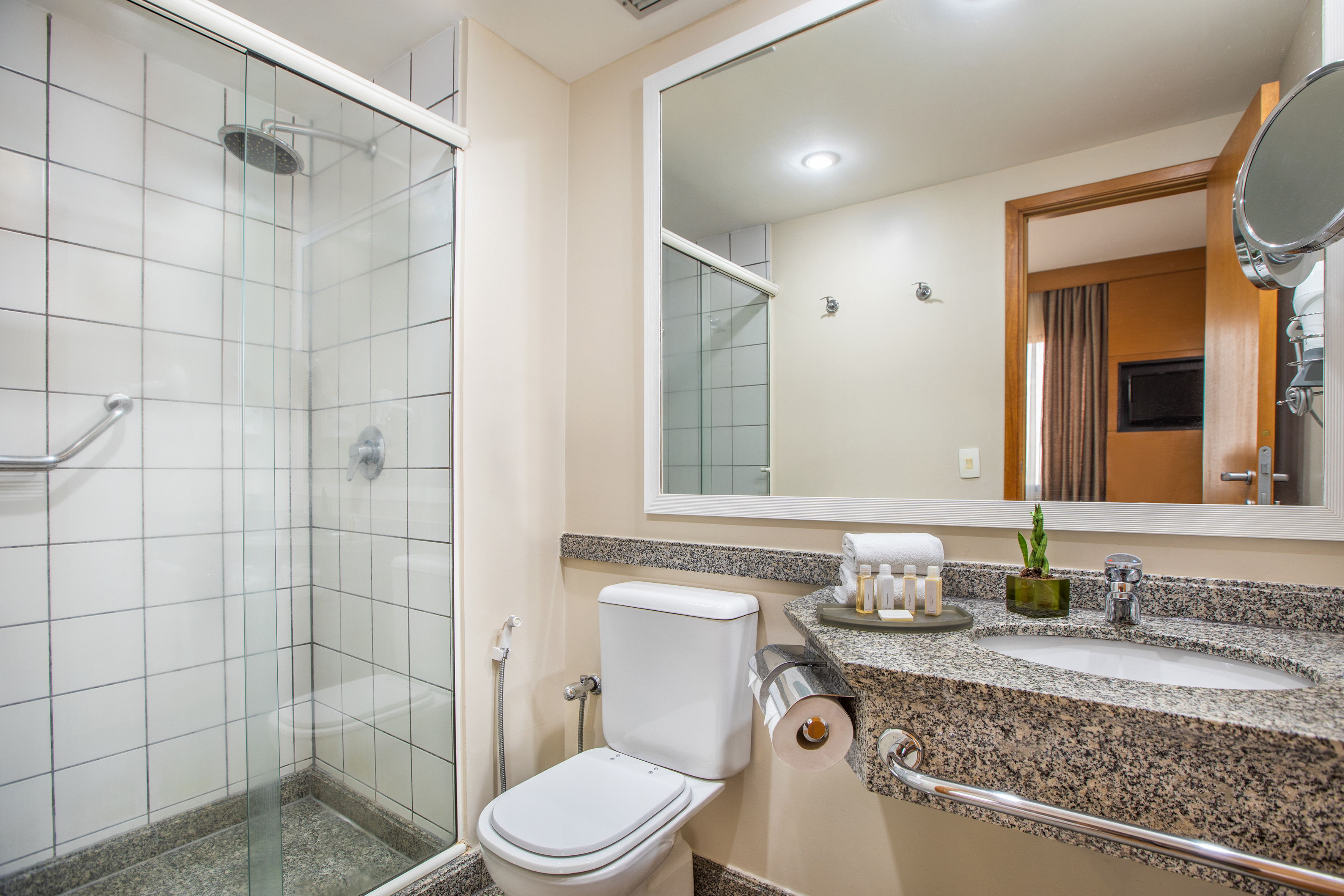 Modern hotel bathroom with granite vanity, glass shower, toilet, amenities.