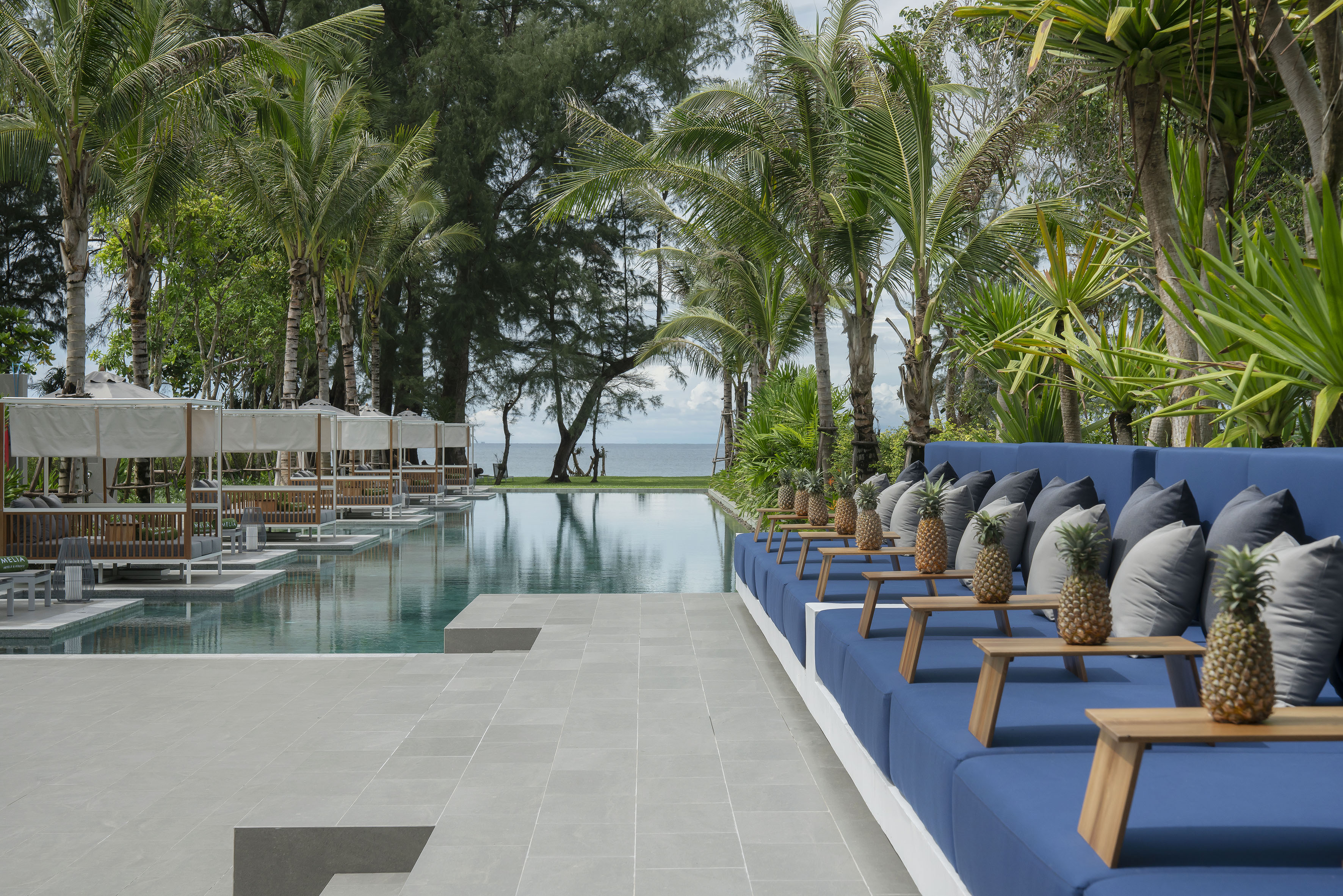 Tropical resort infinity pool with ocean view, palm trees, blue daybeds, cabanas.