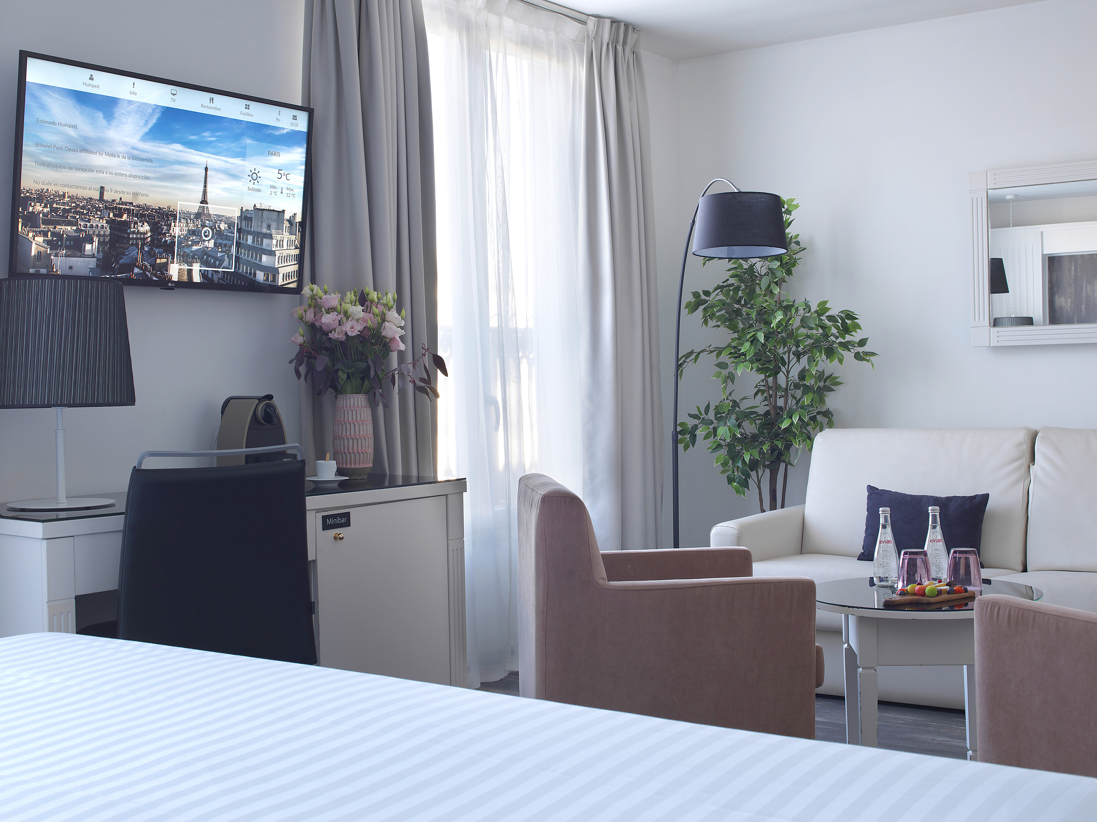 Hotel suite with TV displaying Paris, sofa, armchairs, and minibar.