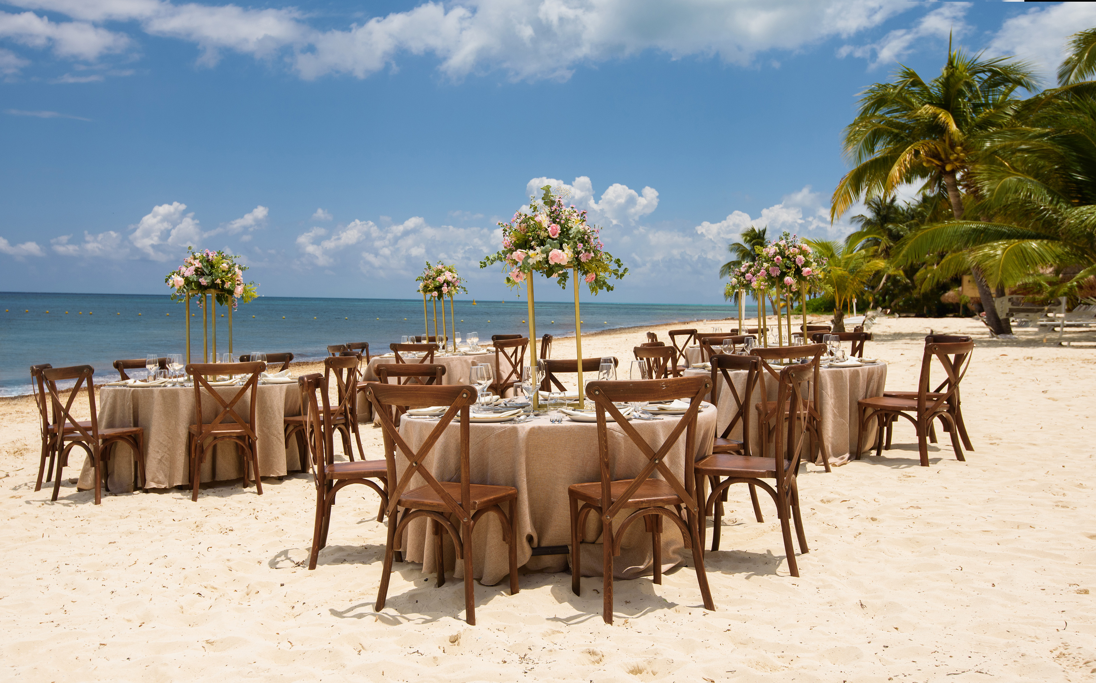 Beach wedding reception with tables, cross-back chairs, floral centerpieces, ocean, palm trees.