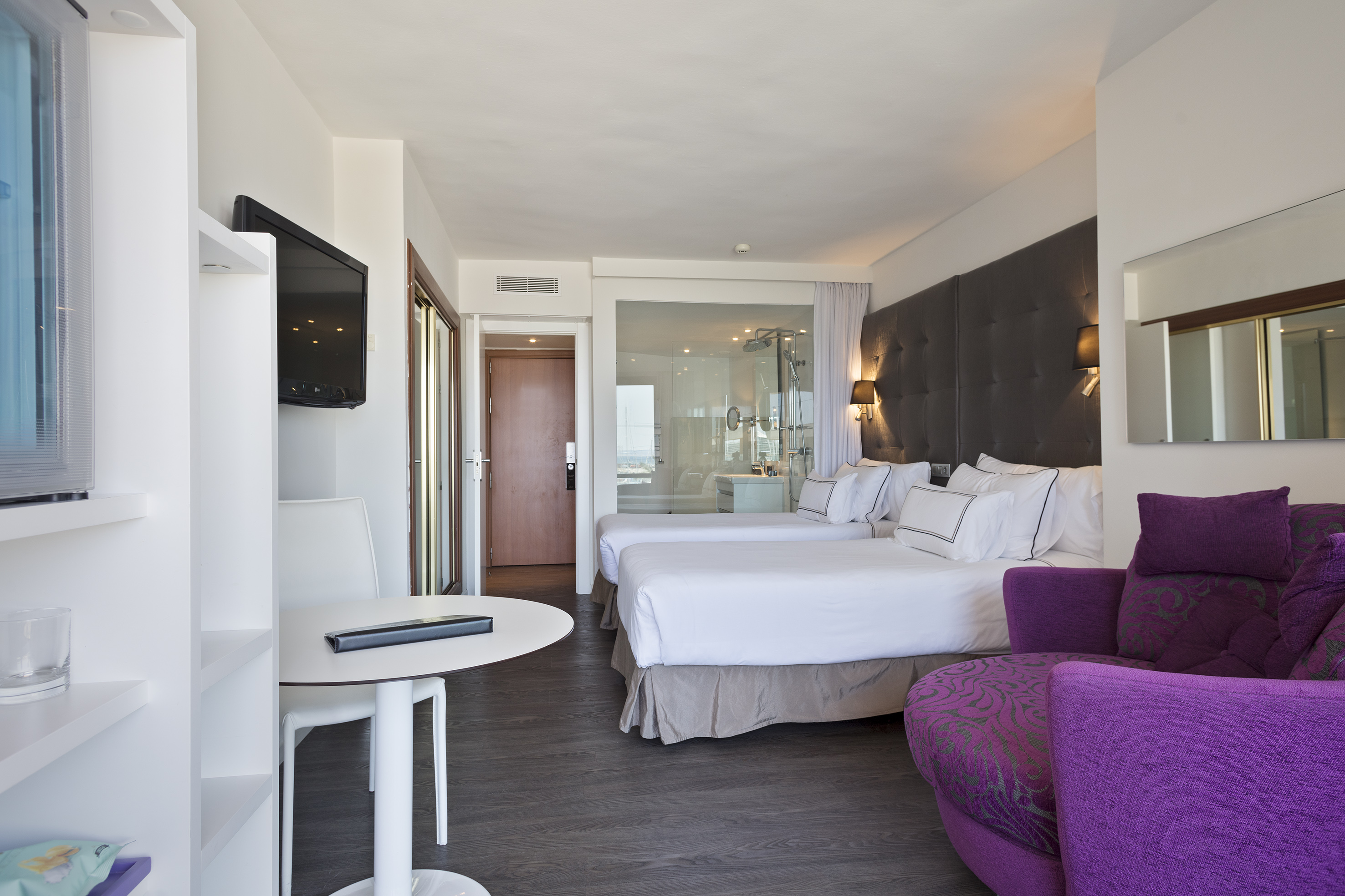Modern hotel room with twin beds, purple sofa, flat-screen TV, and glass shower.