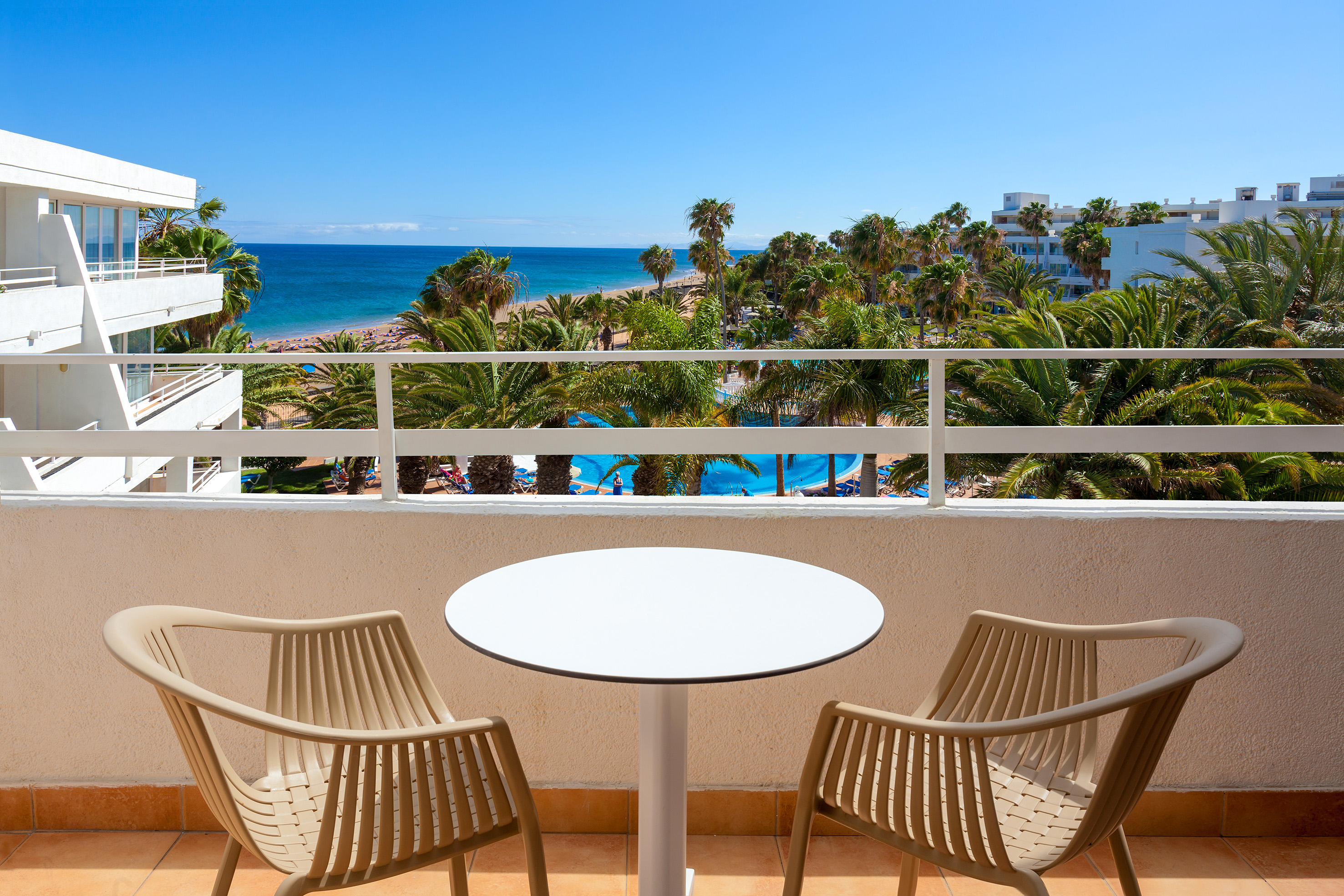 Balcony with table, chairs, ocean view, sandy beach, palm trees, and hotel pool.