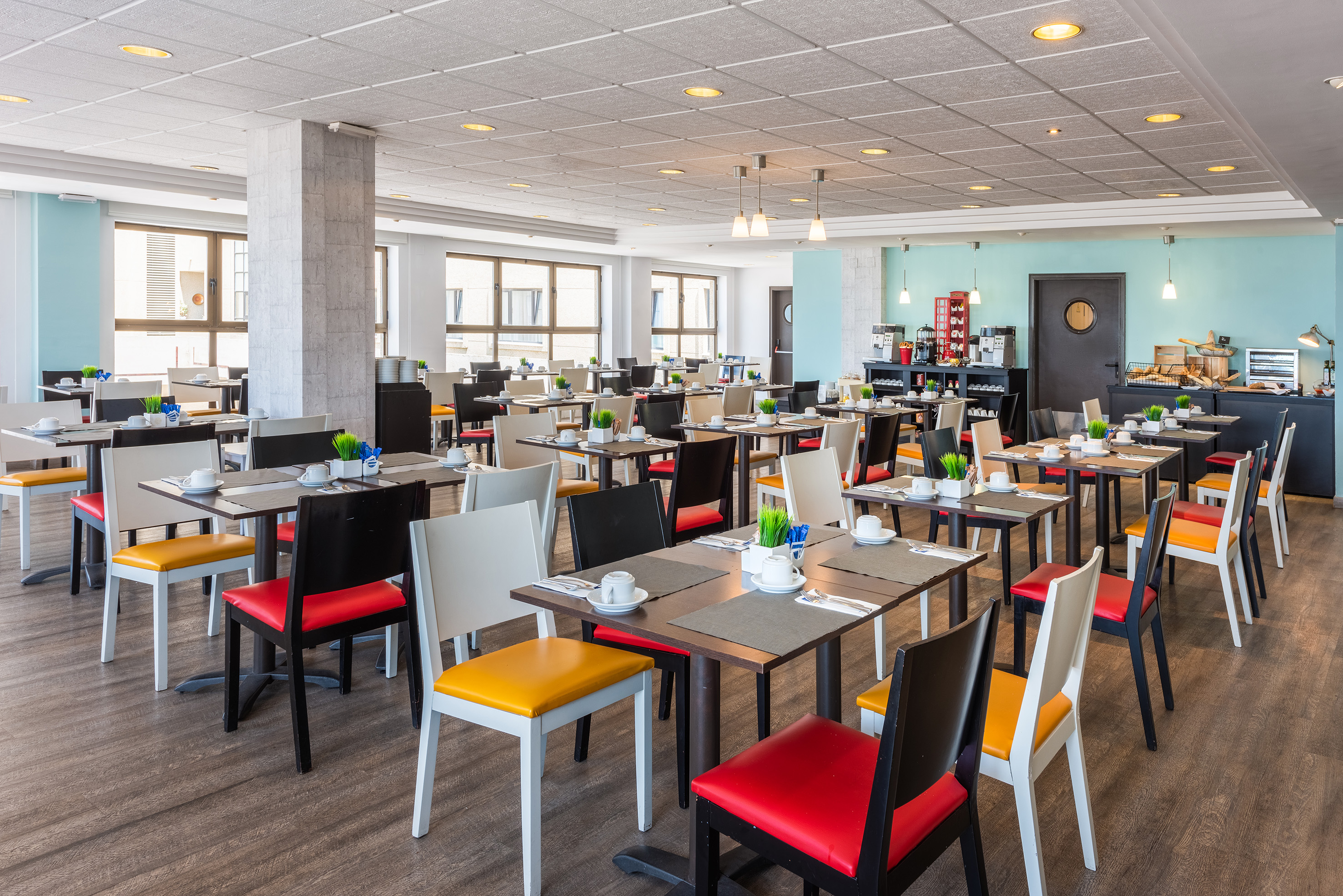 Modern hotel dining room with set tables, colorful chairs, and breakfast buffet.