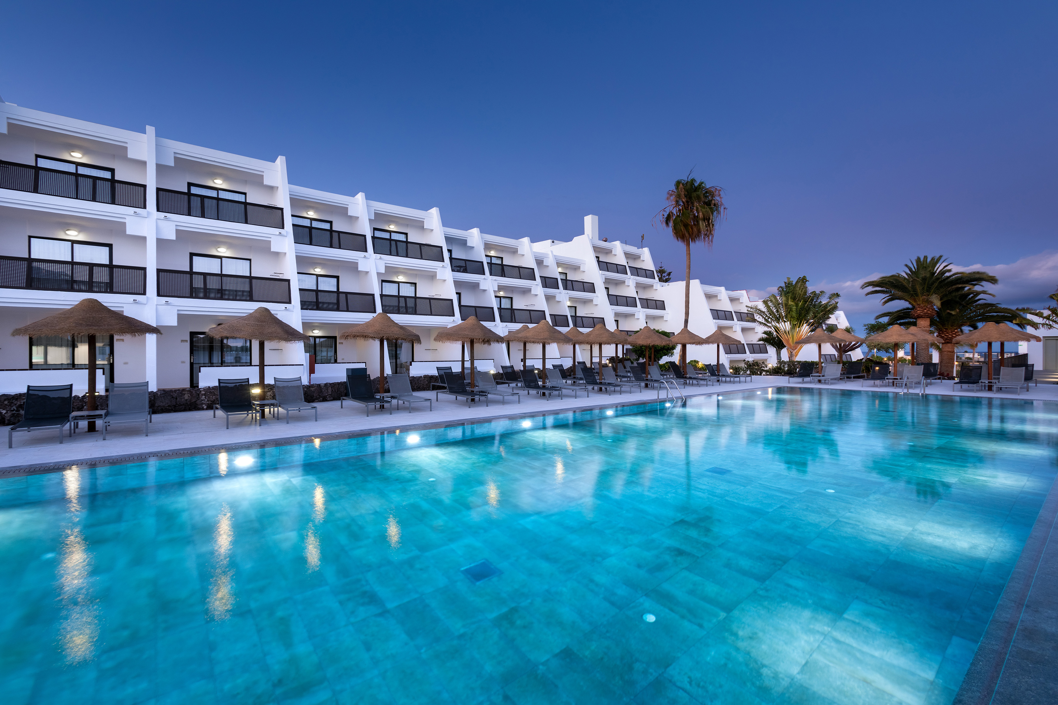 Resort hotel pool with illuminated water, palm trees, and loungers at dusk.