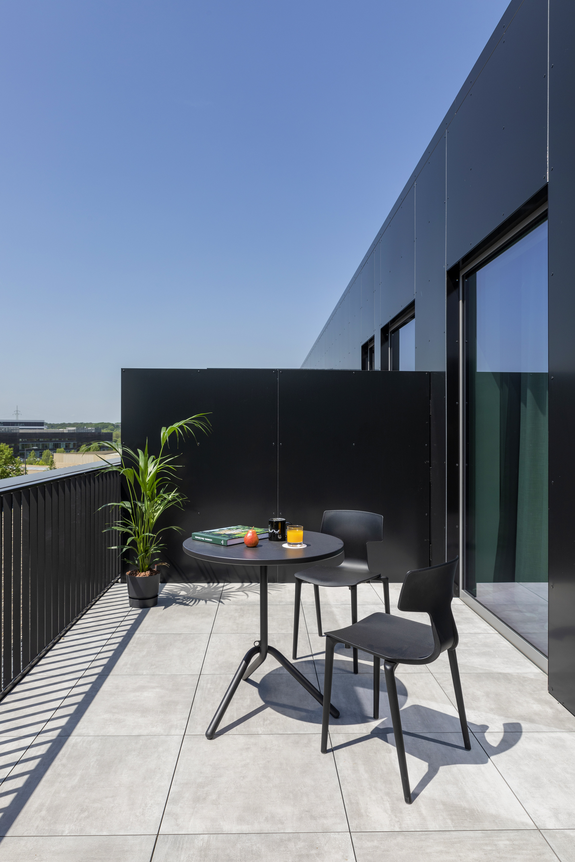 Sunny modern balcony with black outdoor table, chairs, plant, and tiled floor.