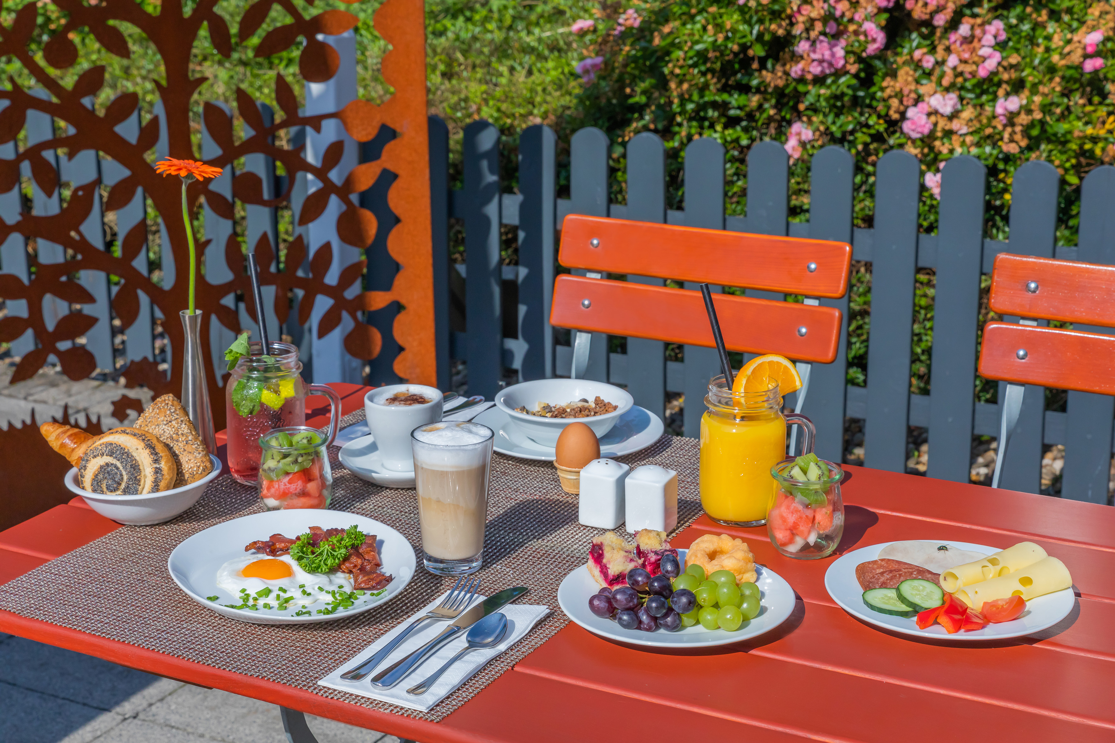 Outdoor breakfast on red table with fried egg, bacon, fruit, coffee, and rolls.