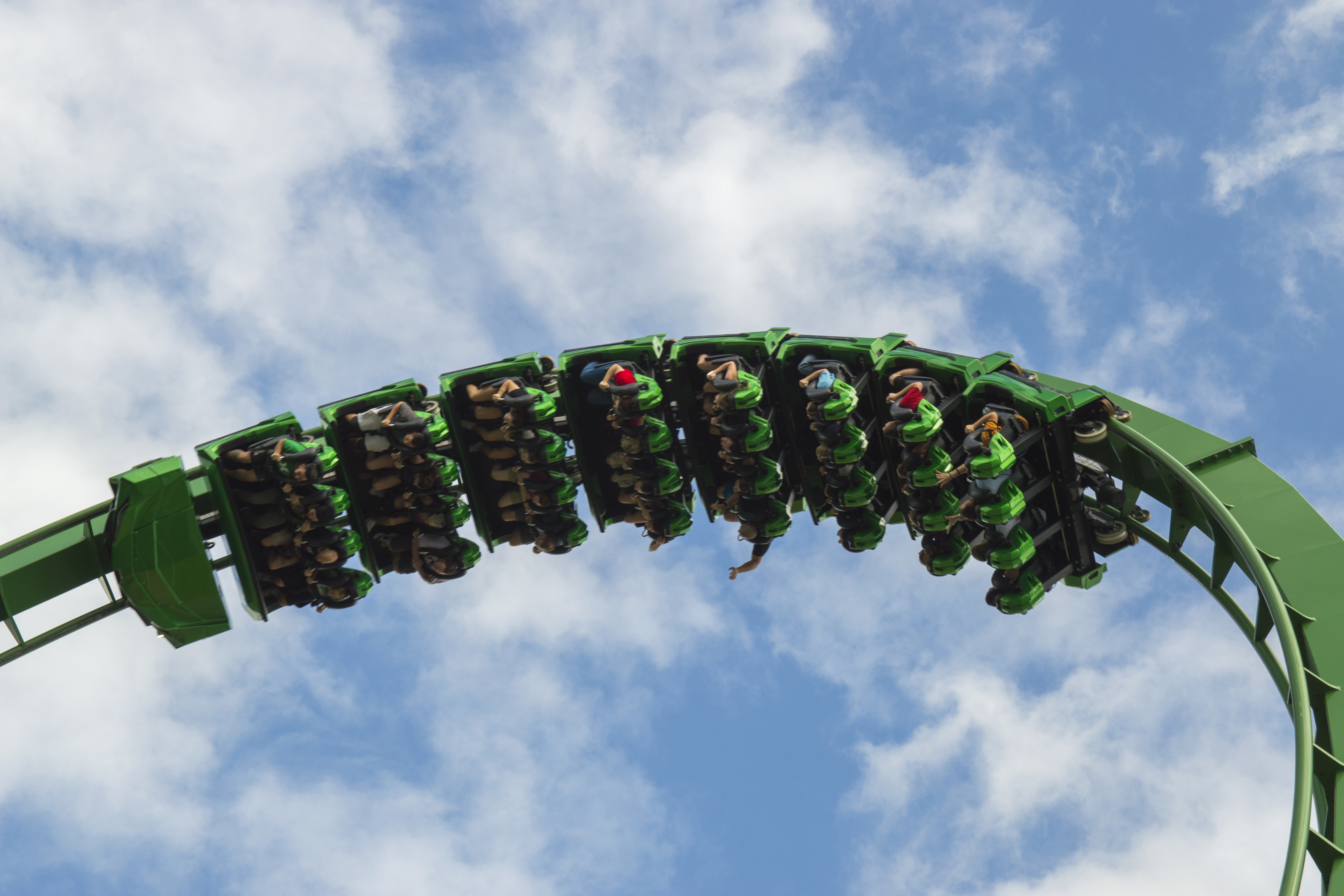 Green inverted rollercoaster with riders upside down against a cloudy blue sky. - Universal Epic Universe™ - Meliá Orlando Celebration, Orlando