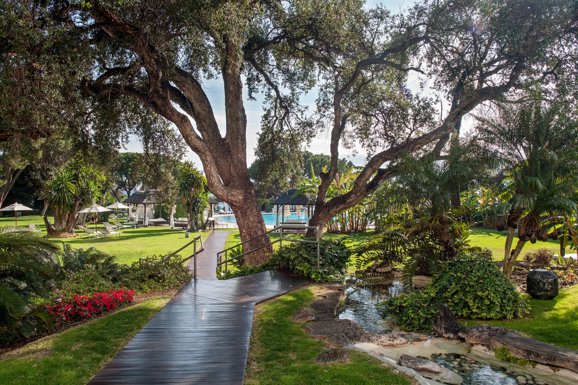 Luxury resort garden path leading to pool, surrounded by trees and stream.
