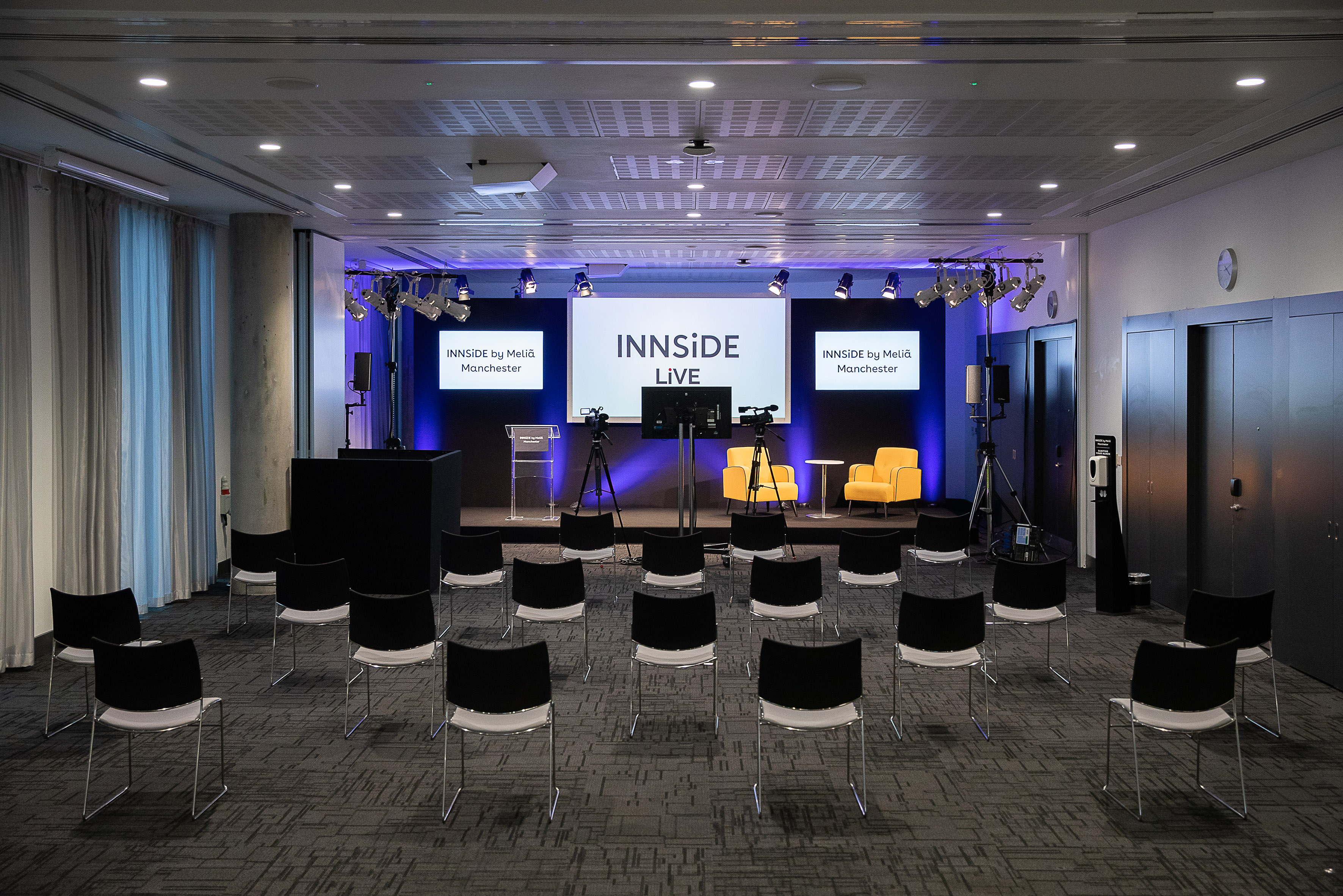 INNSiDE Manchester conference room with stage, screens, cameras, and audience seating.