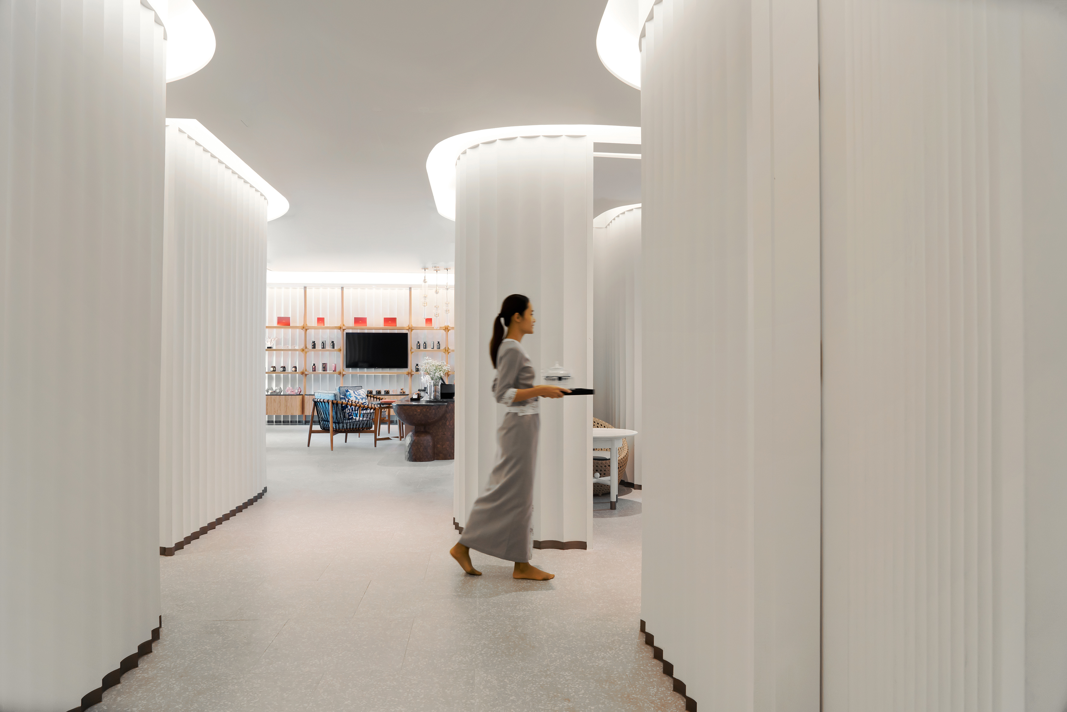 Barefoot woman carries tray in modern spa/wellness center with curved white walls.