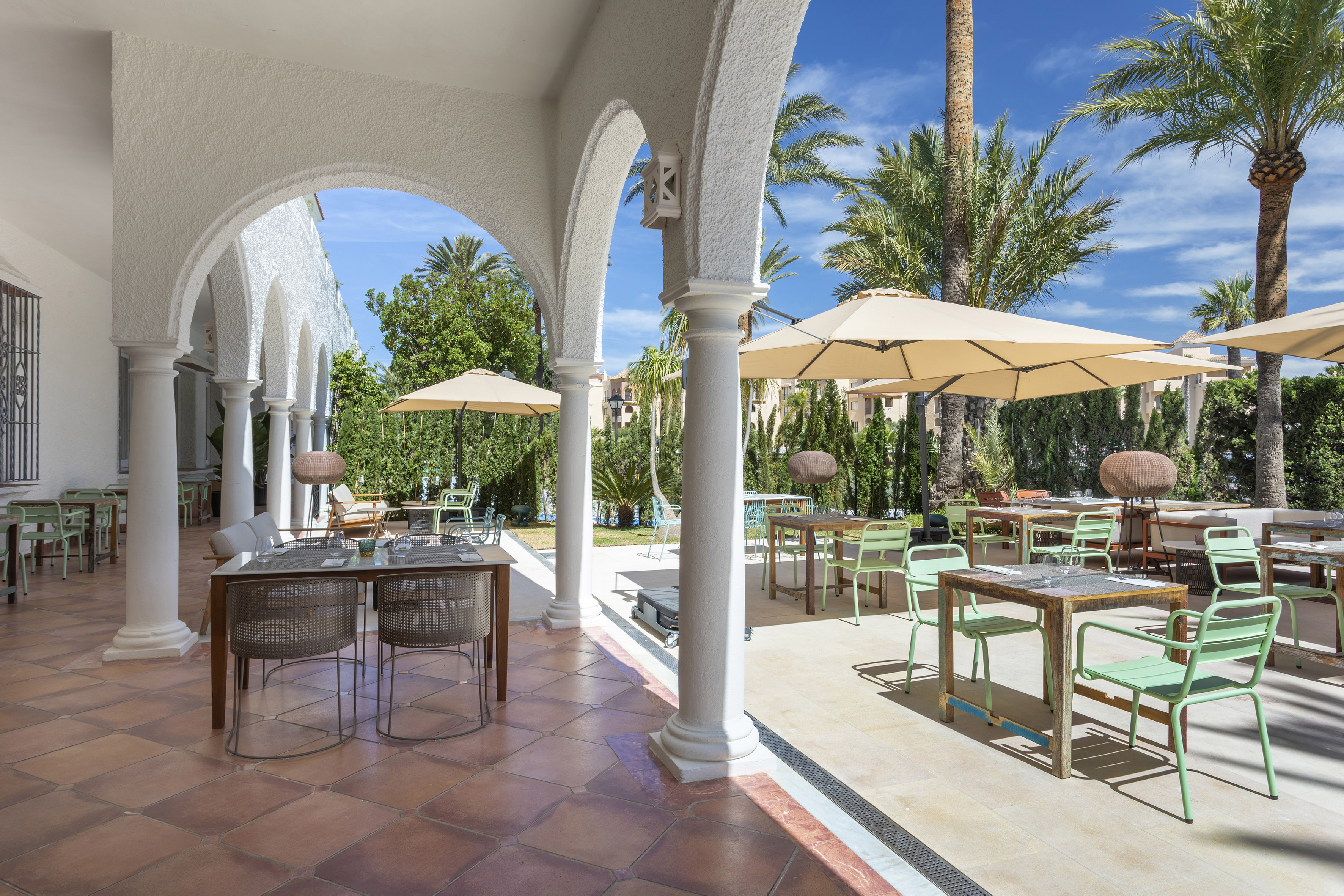 Outdoor restaurant terrace with white arches, shaded tables, palm trees.