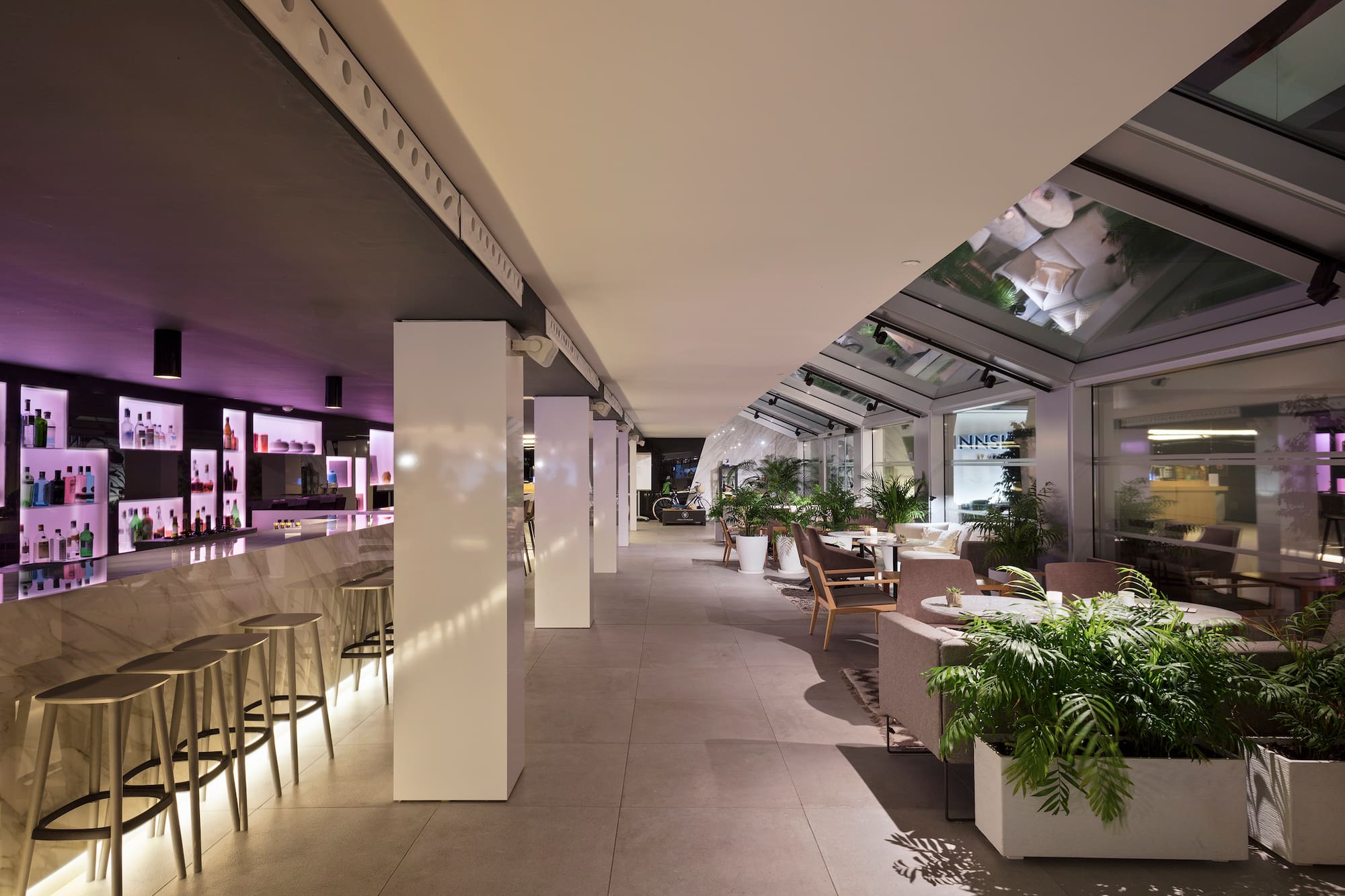 Stylish hotel bar with glowing counter, lounge seating, and indoor plants.