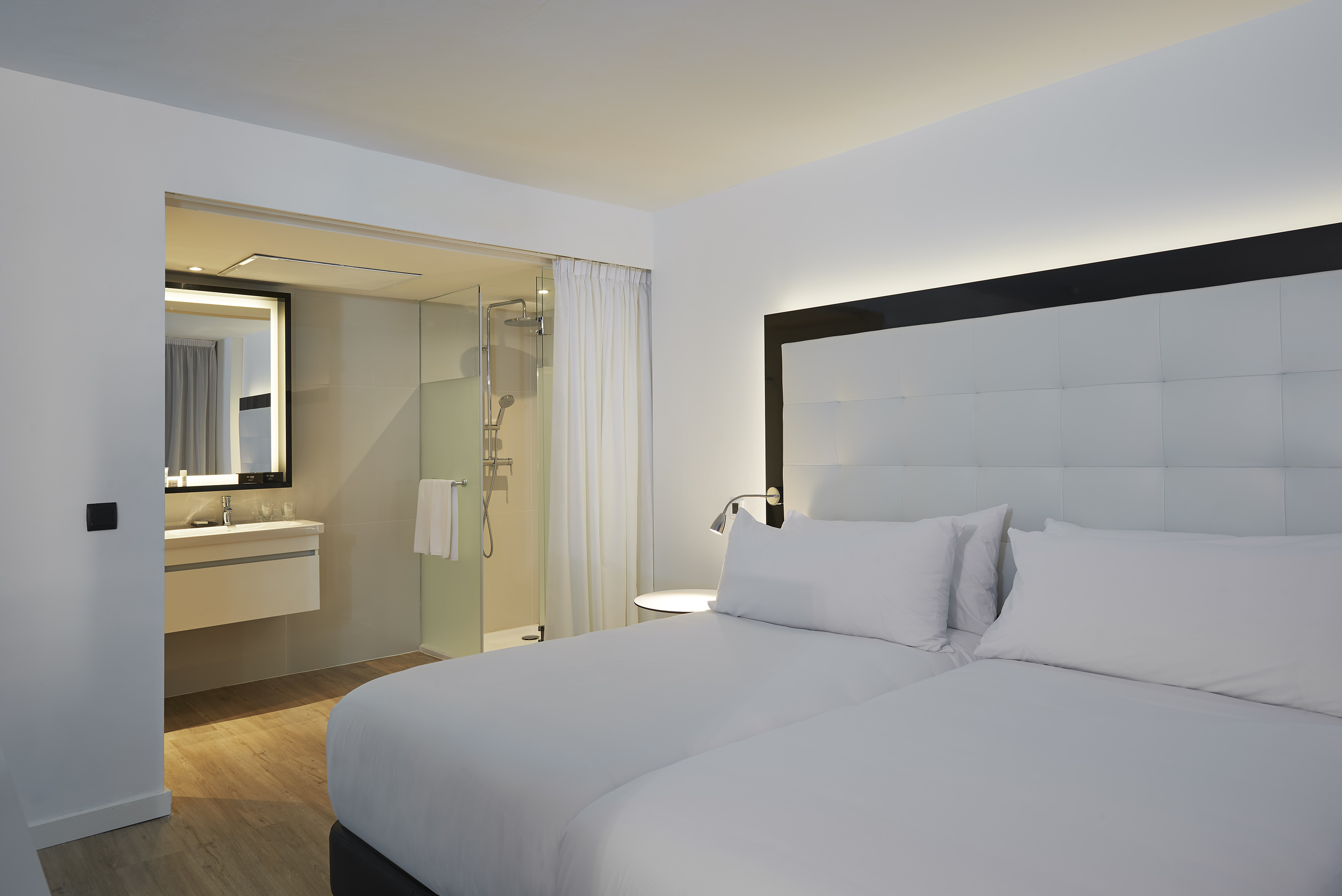 Modern hotel room with twin beds, white linens, and integrated glass shower bathroom.