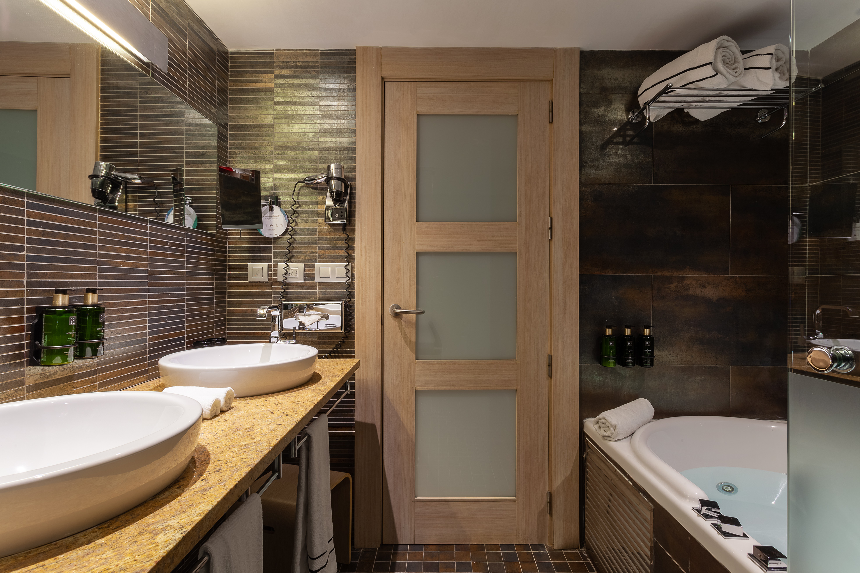 Modern hotel bathroom with double vanity, vessel sinks, brown tiles, and a bathtub.