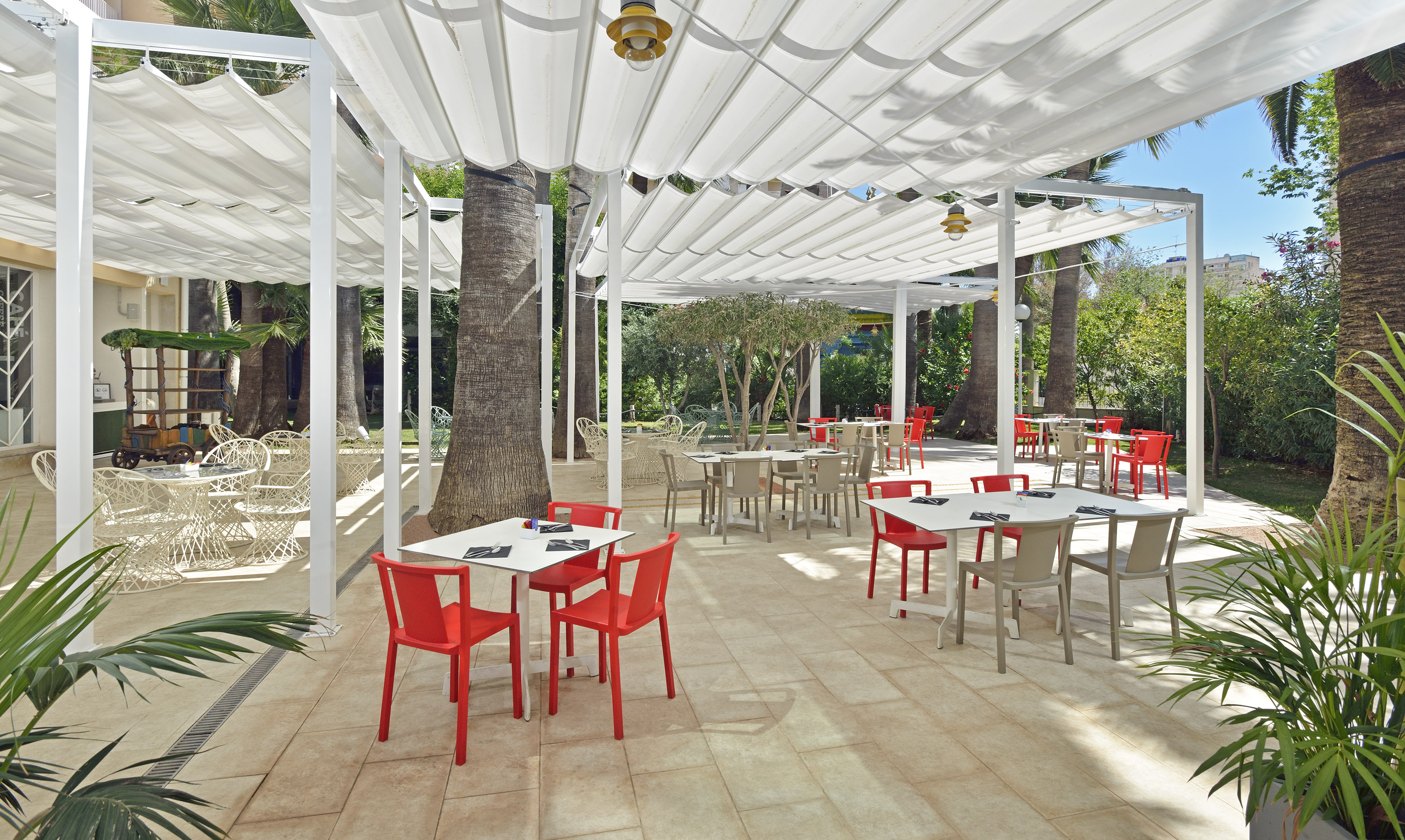Resort outdoor dining patio with white pergola, red chairs, and palm trees.
