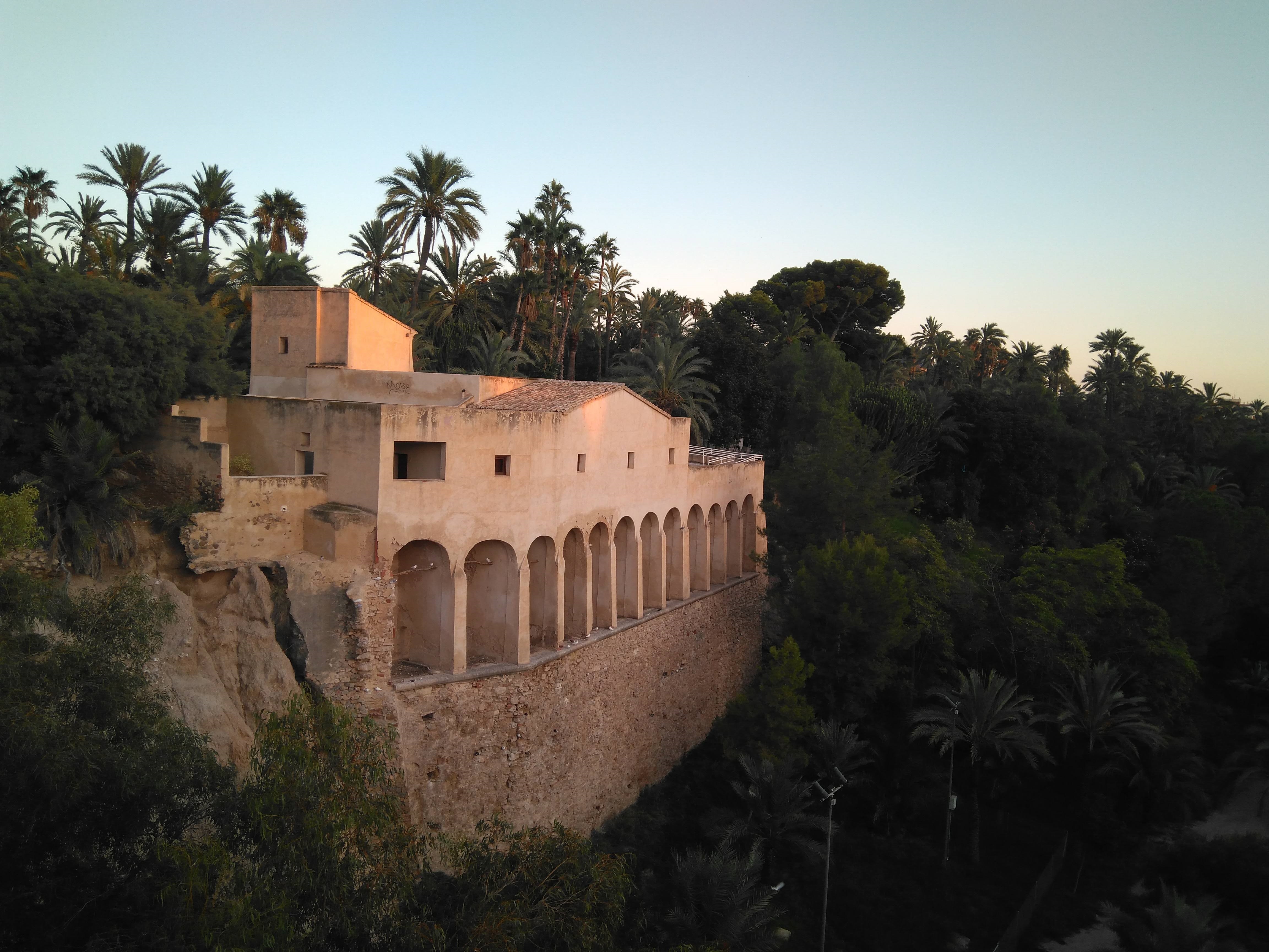 Historic arched building on cliffside with dense palm groves at sunset.