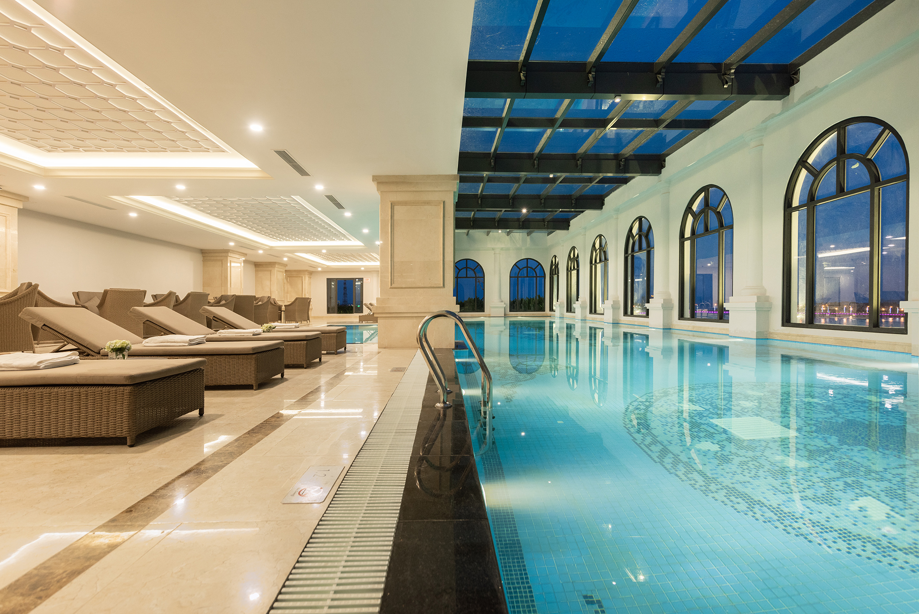 Elegant indoor swimming pool with lounge chairs, arched windows, and a glass skylight.